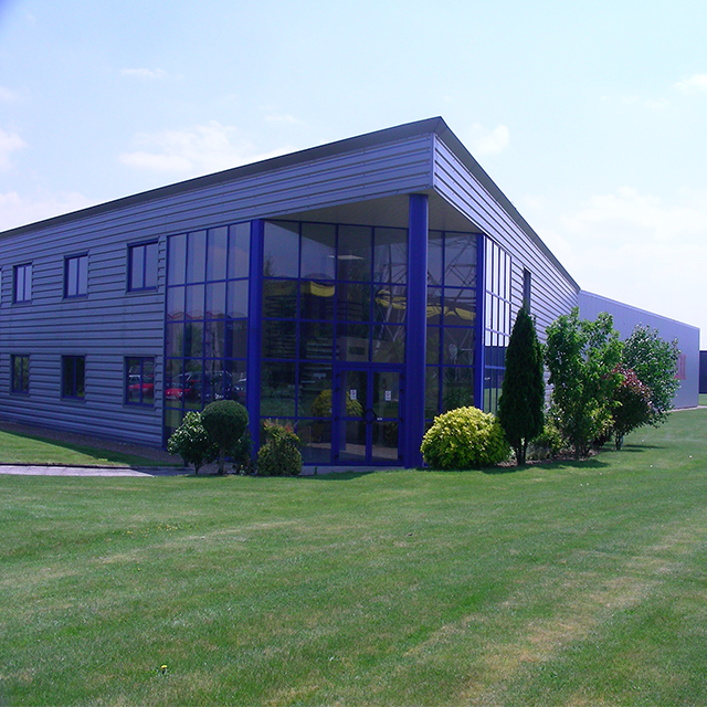 Modern office building with blue glass and metal facade, surrounded by neatly trimmed green lawn and bushes.