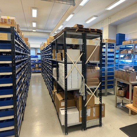 A warehouse aisle with metal shelves filled with cardboard boxes and organized items. The room is well-lit, tidy, and has a functional atmosphere.