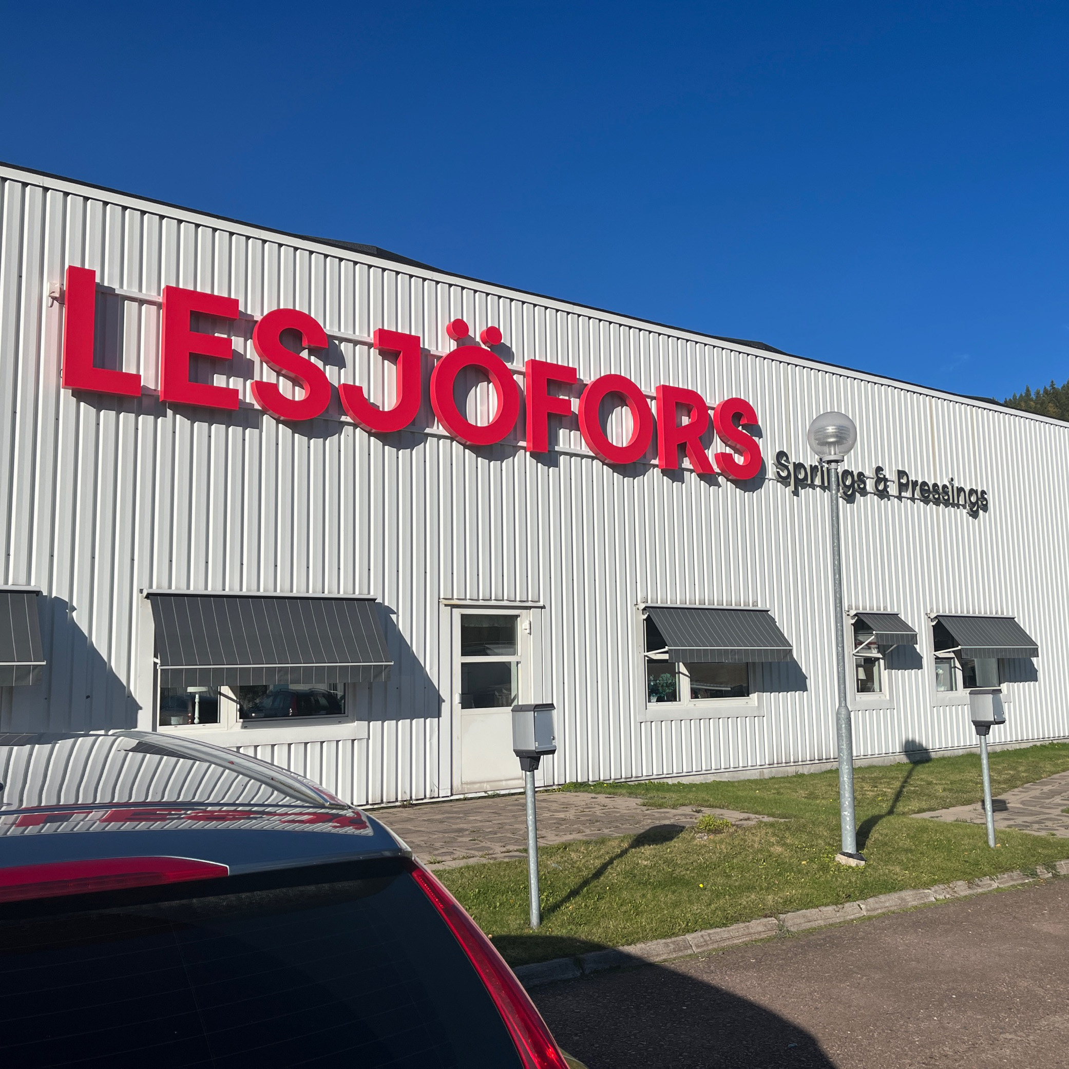 White industrial building with large red "LESJÖFORS" sign and smaller text reading "Springs & Pressings." Clear blue sky and trees in the background.