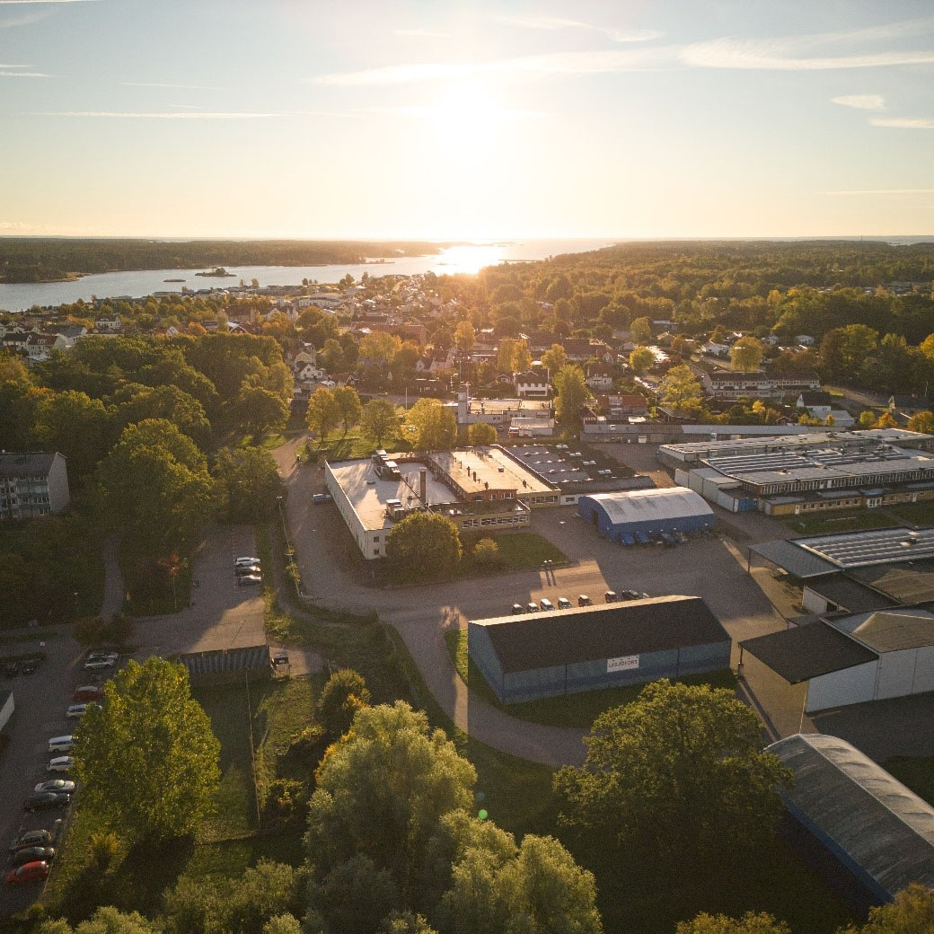 A serene aerial view of a small town at sunset, featuring green trees, clustered buildings, and a distant shimmering body of water, evoking tranquility.