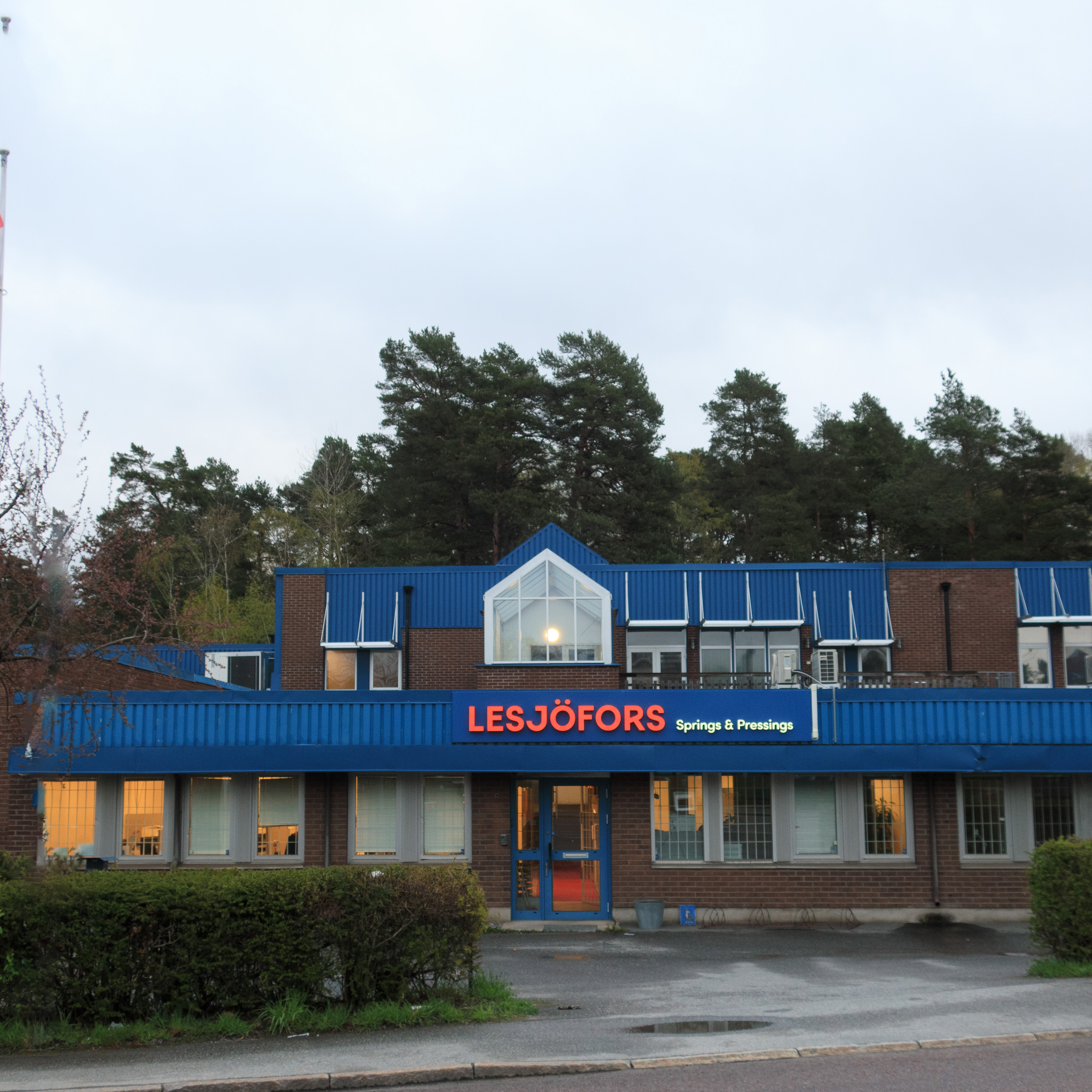 Brick building with a blue roof and "Lesjöfors Springs & Pressings" signage. Surrounded by trees, creating a calm and industrial atmosphere.
