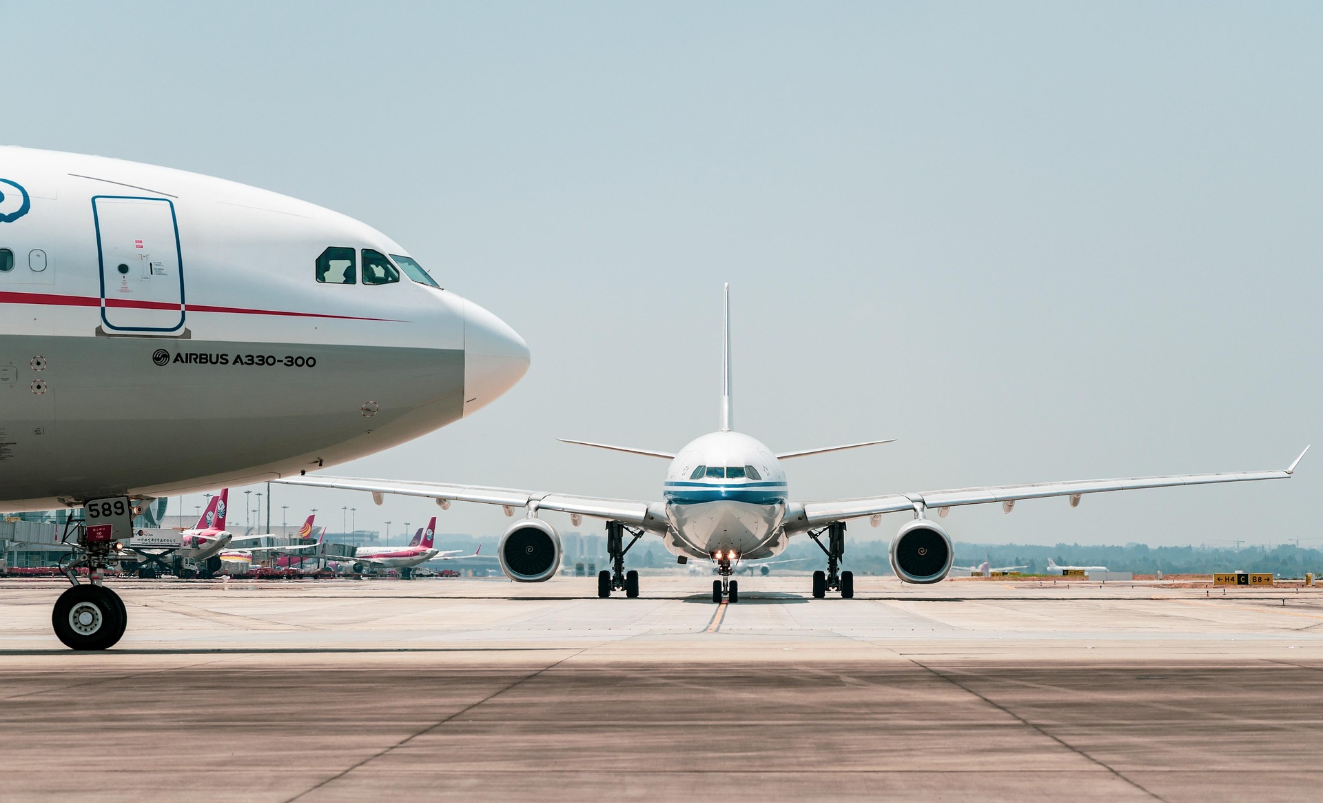 Front view of a commercial airplane taxiing on a runway, with a large Airbus aircraft parked to the left and additional planes in the background.