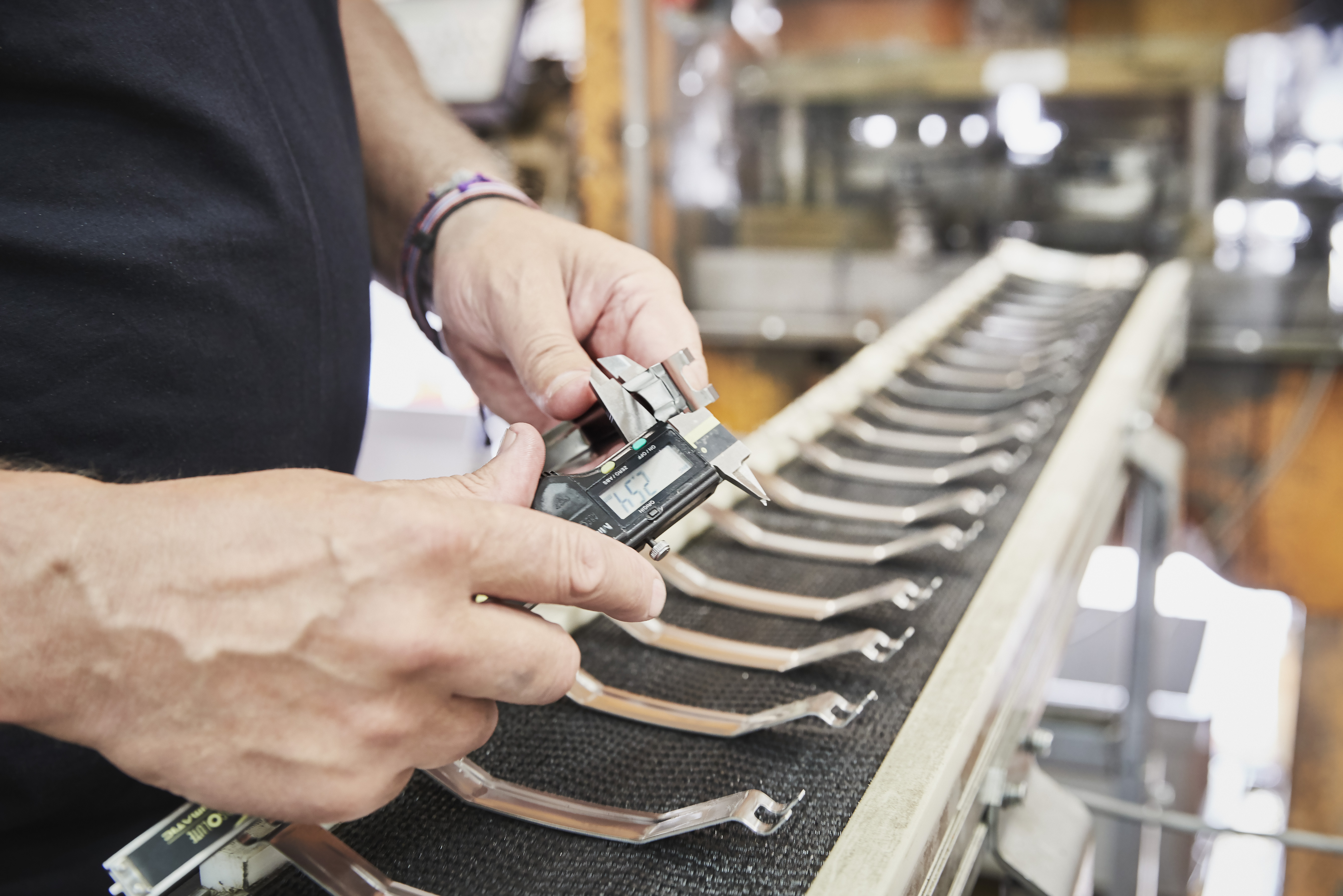 A worker using digital calipers to measure a formed metal part, with multiple identical components lined up on a conveyor belt.