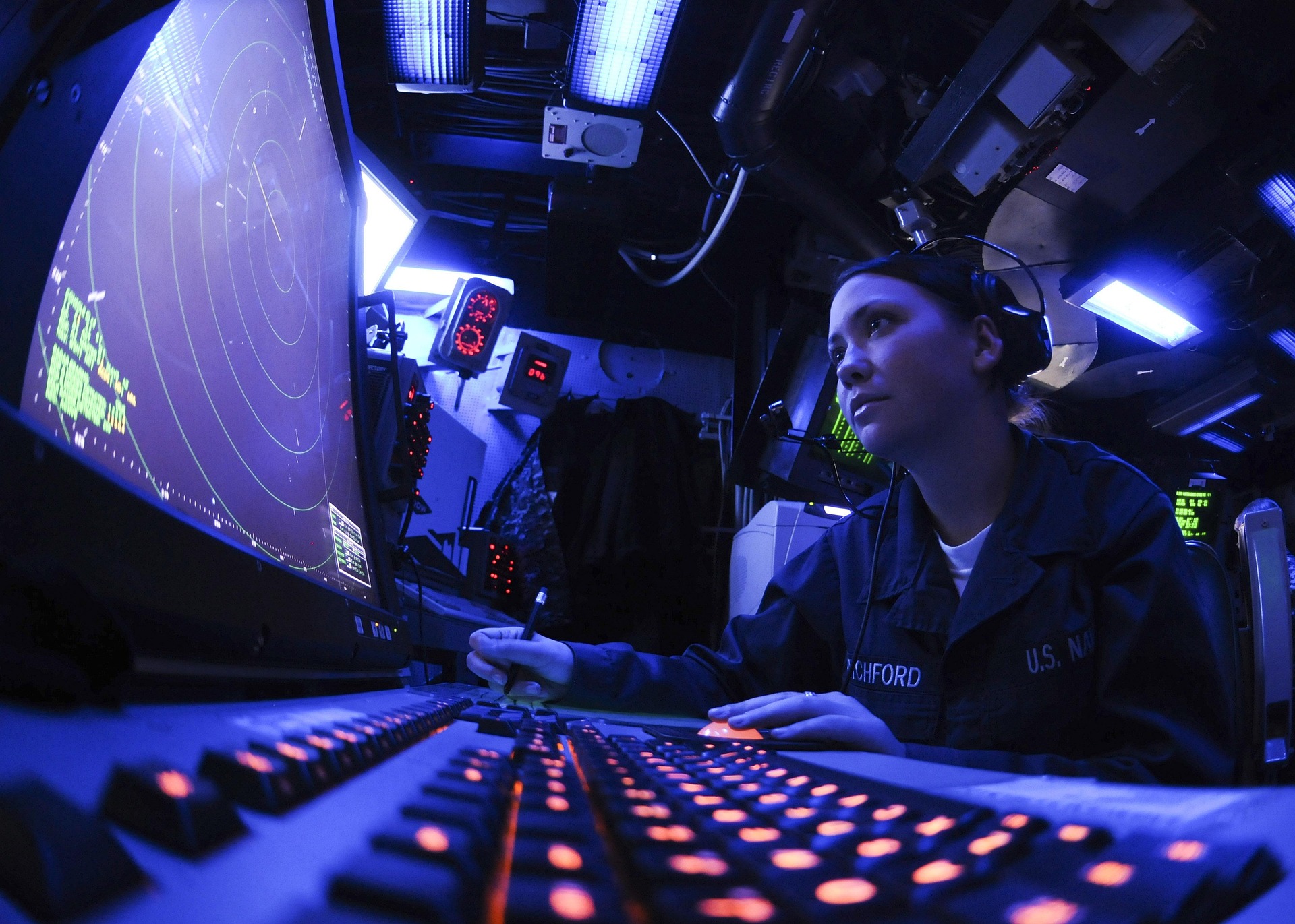 A Navy officer wearing a headset operating a radar system in a dark control room.