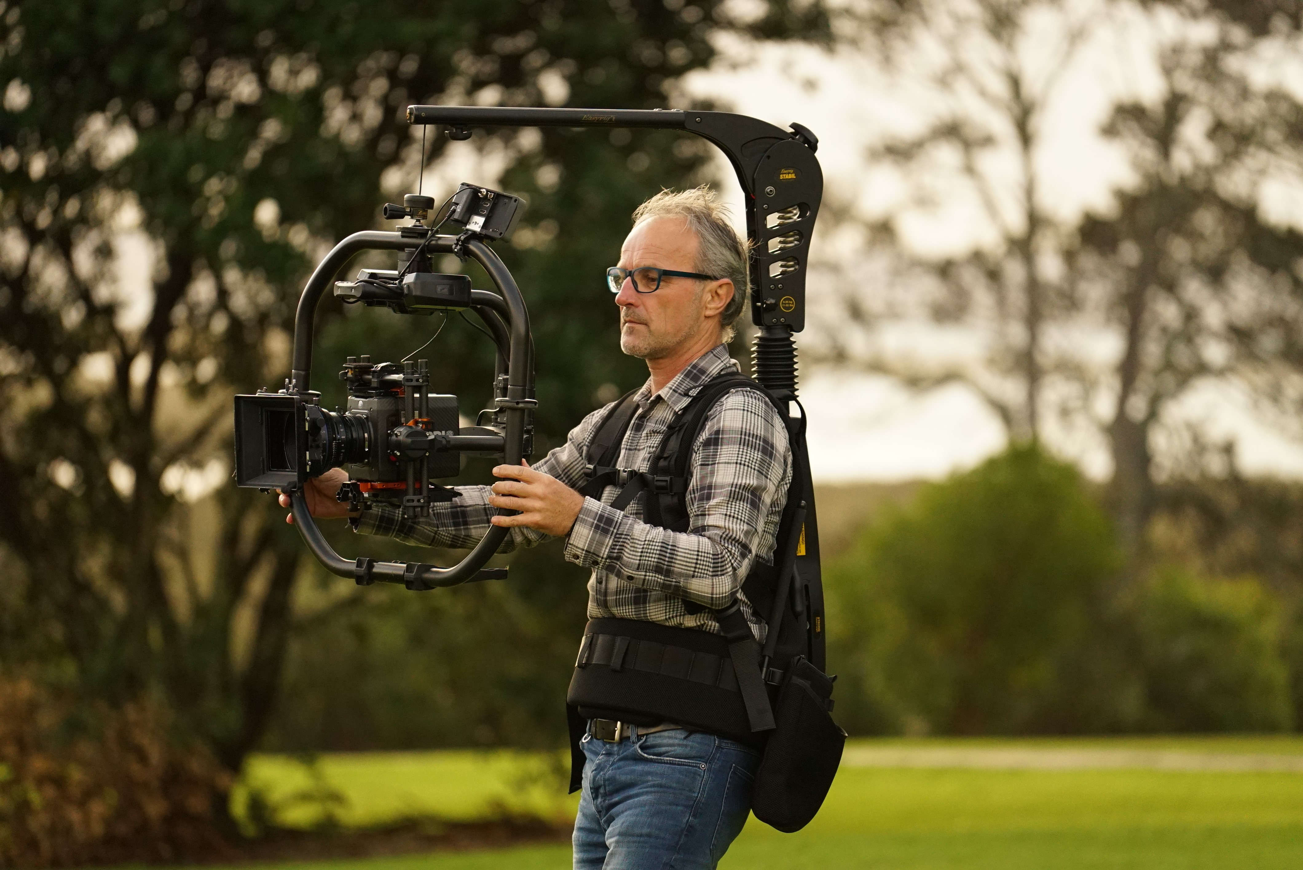 A person wearing a plaid shirt operates a camera stabilizer rig in a lush park setting, focused and professional, capturing outdoor scenes.