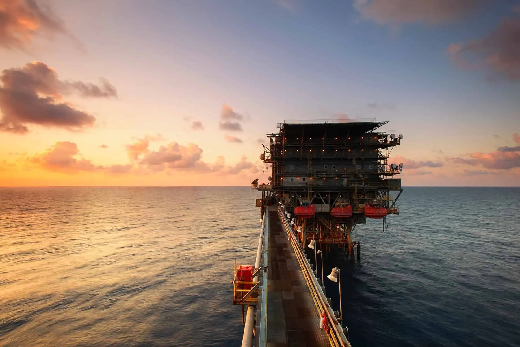 Offshore energy platform viewed from a walkway, set against the ocean at sunset.