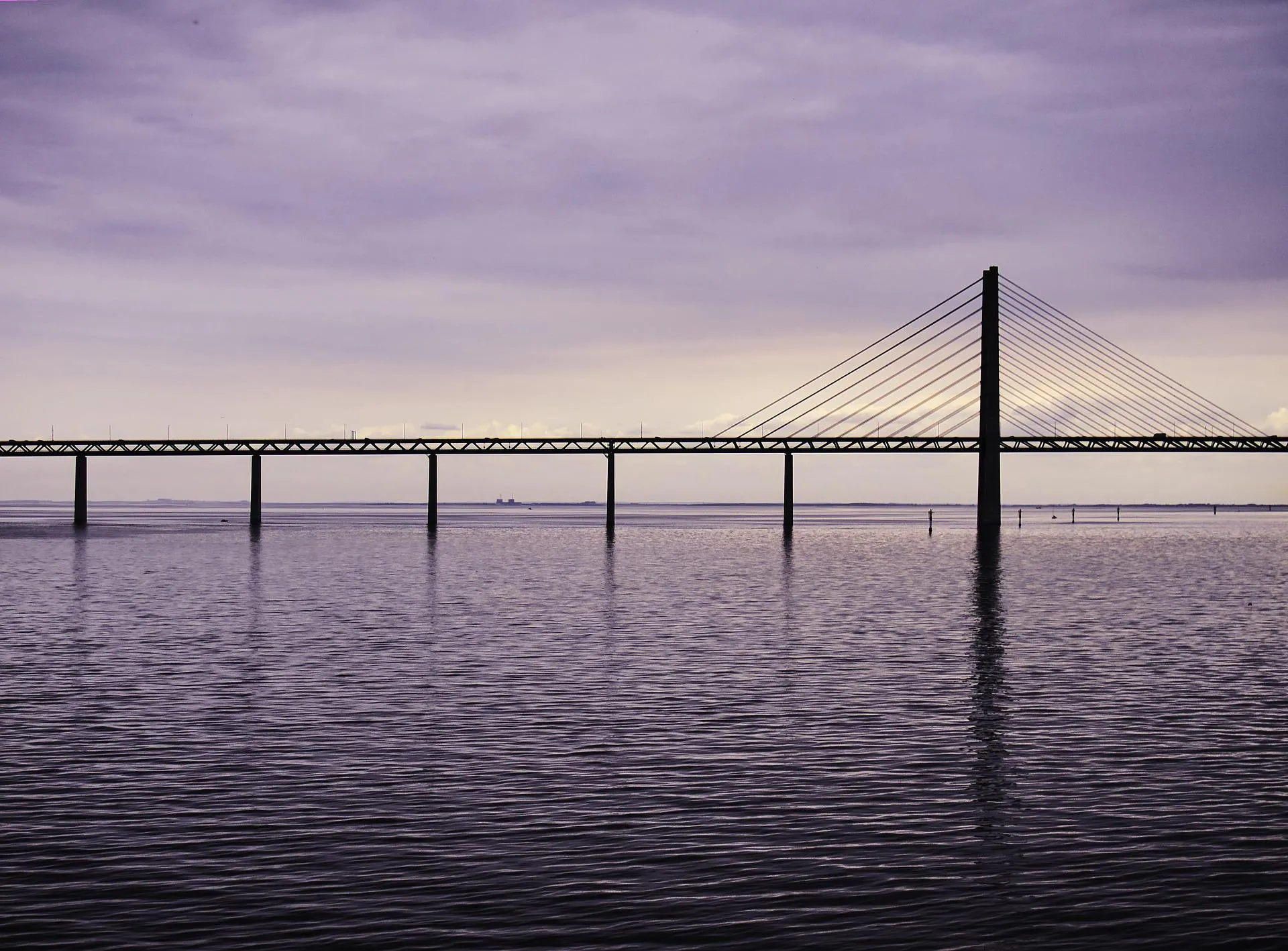 The Øresund Bridge silhouetted against a moody purple sky, reflected in calm water, with one of the cable-stayed pylons visible on the right.