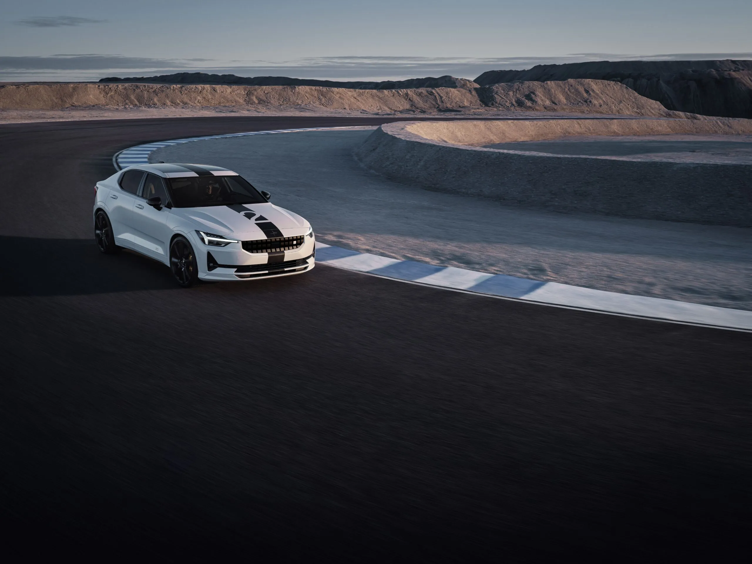 A white Polestar 2 BST Edition with black racing stripes navigating a corner on a race track, shot from an elevated angle at dusk."