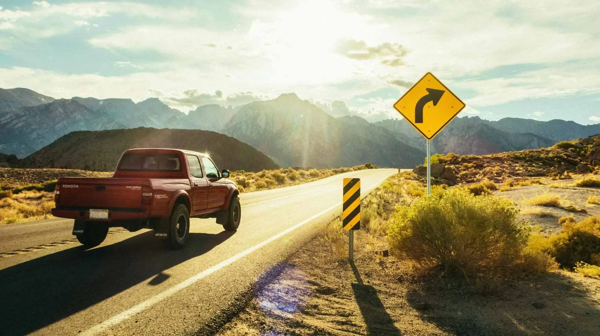 A red pickup truck drives on a sunlit, winding road through a desert landscape, with mountains in the background and a yellow curve sign ahead.