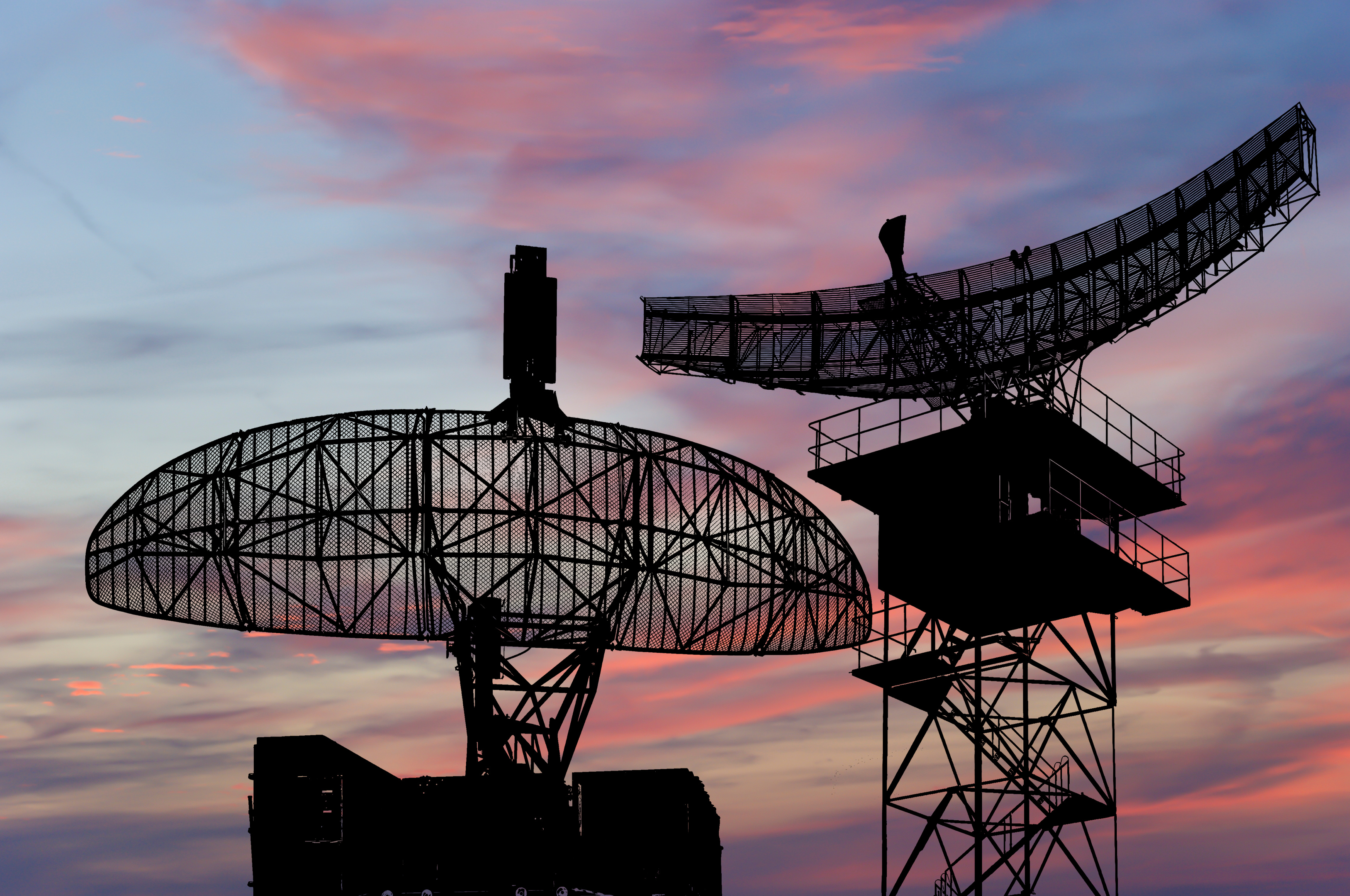 Silhouettes of two large radar antennas against a sunset sky.