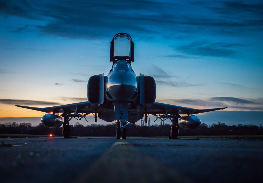 A military jet is parked on a runway at dusk, silhouetted against a vibrant blue and orange sky.