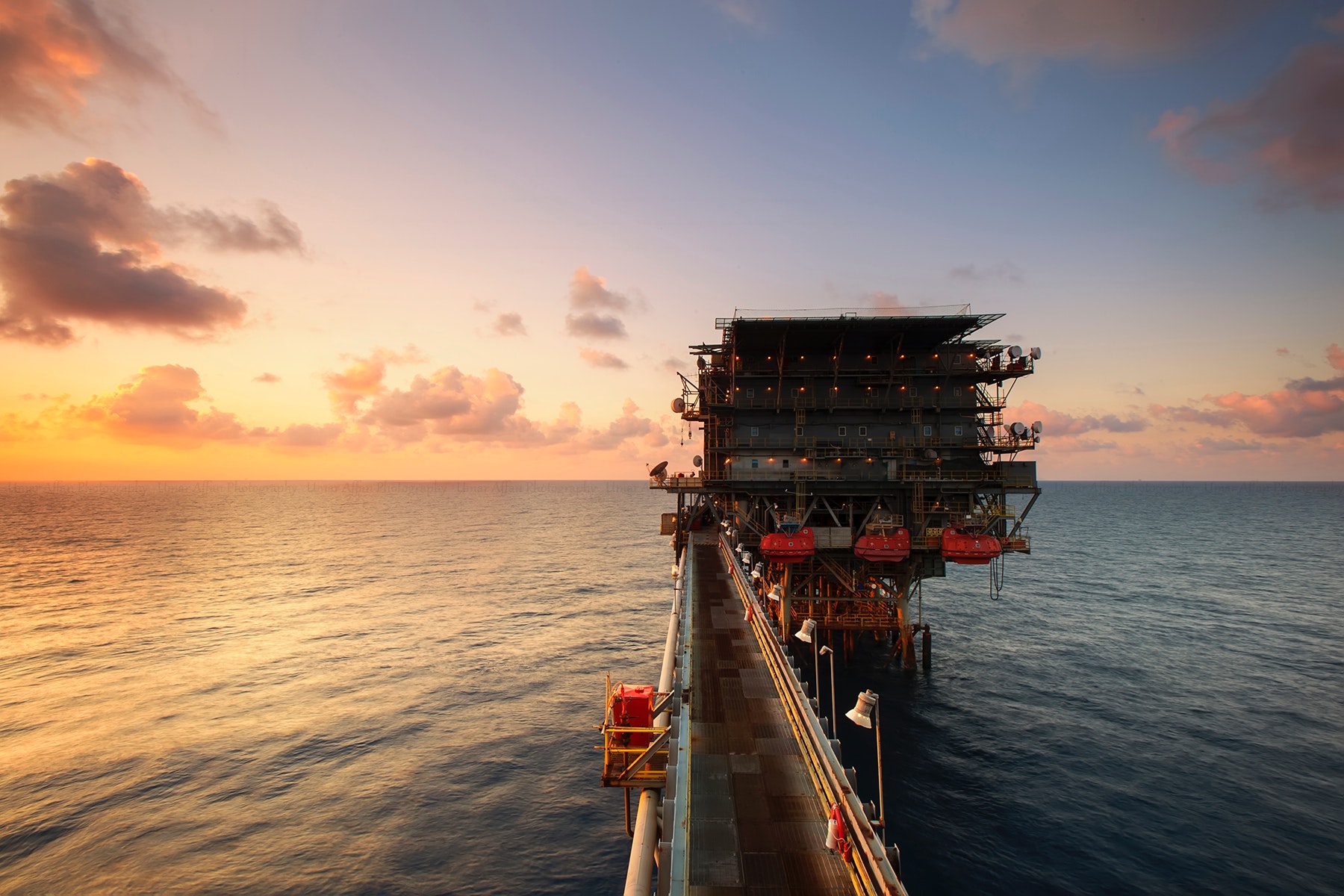 Oil rig extends into a calm ocean under a vibrant sunset sky. The structure appears industrial, with warm colors reflecting a serene yet robust scene.