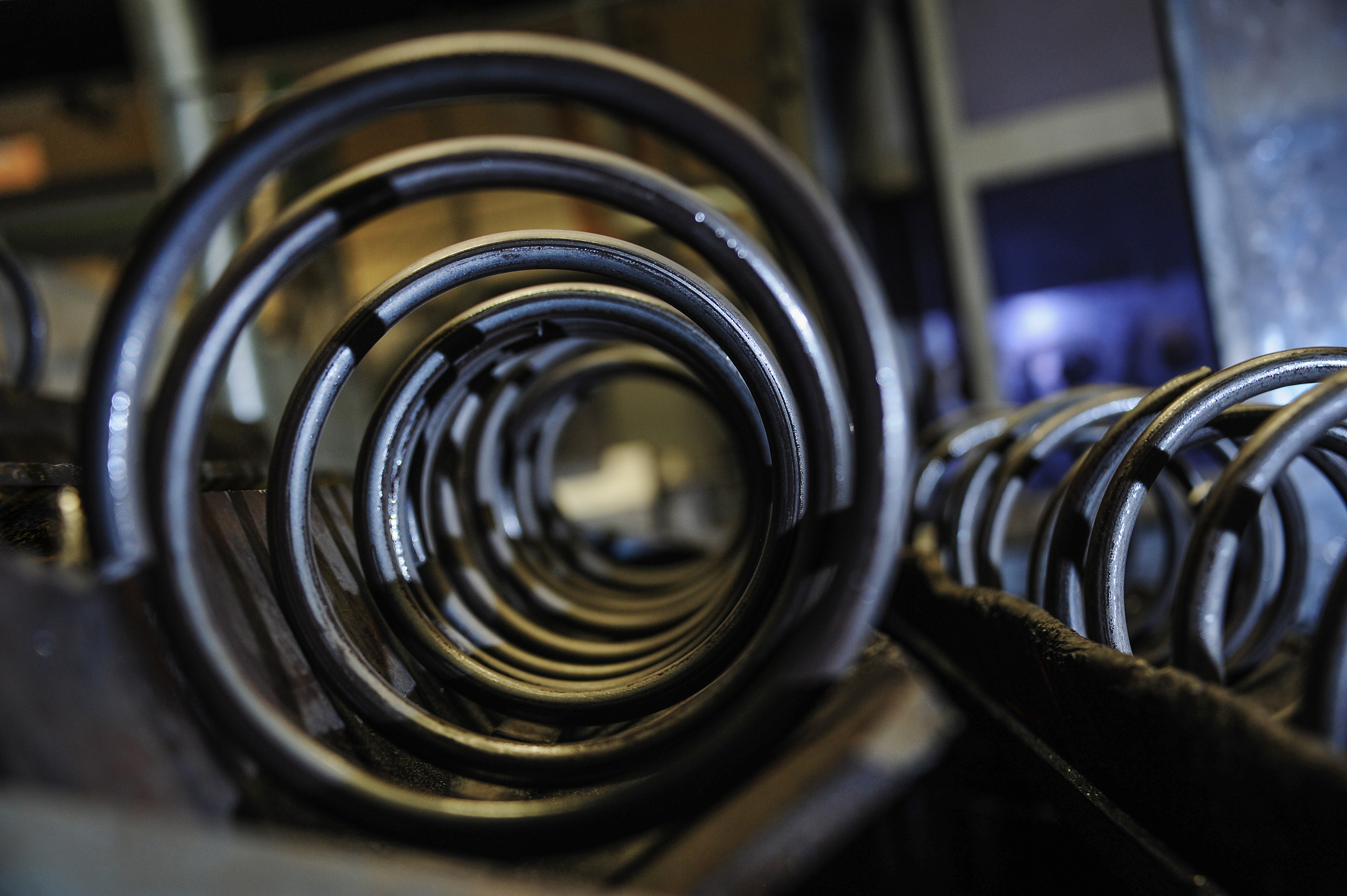 Close-up of stacked coil springs in a dimly lit workshop. The perspective focuses on the spirals, creating a sense of depth and industrial ambiance.