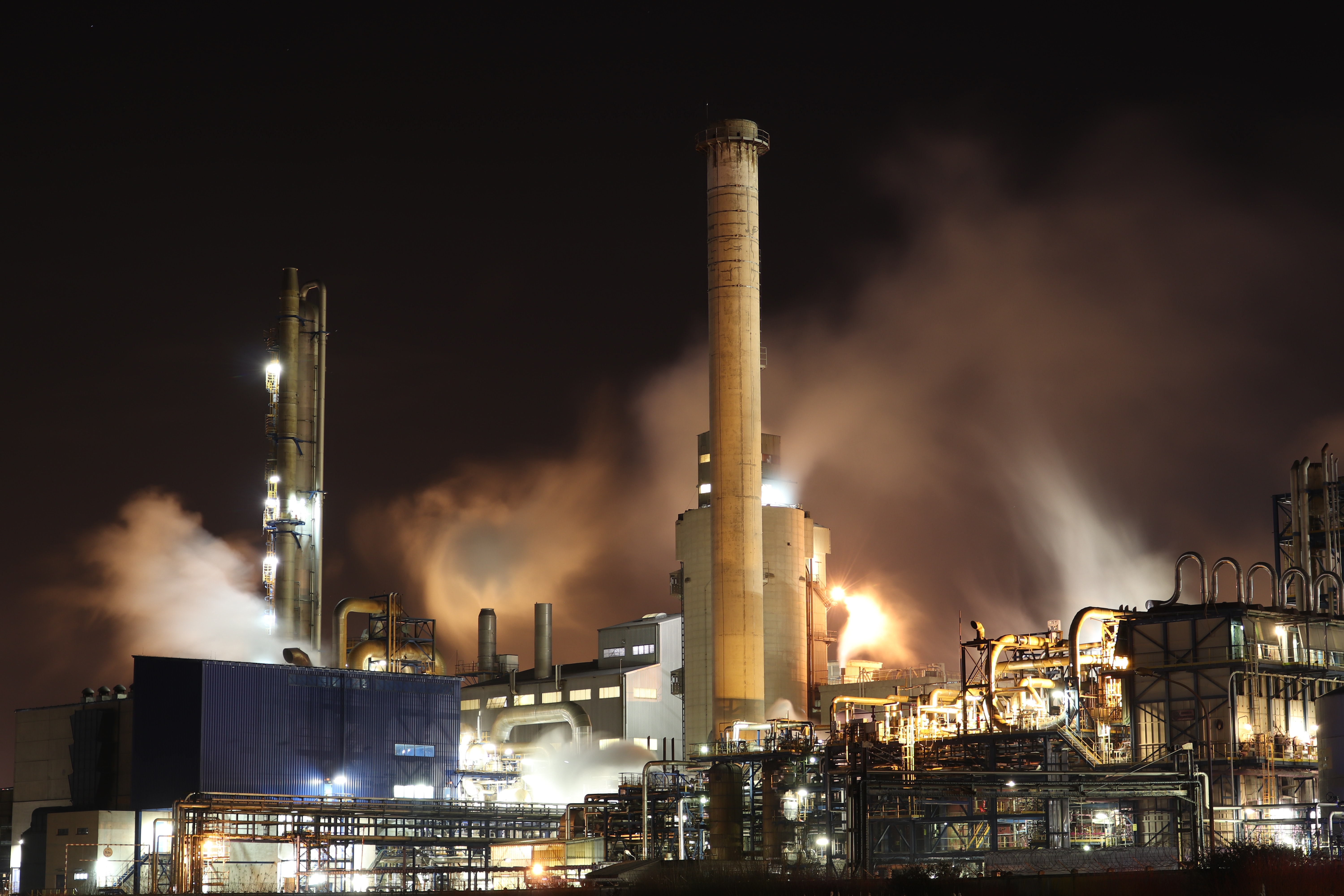 Nighttime industrial scene featuring a factory emitting large plumes of smoke and steam. Bright lights contrast against the dark sky.
