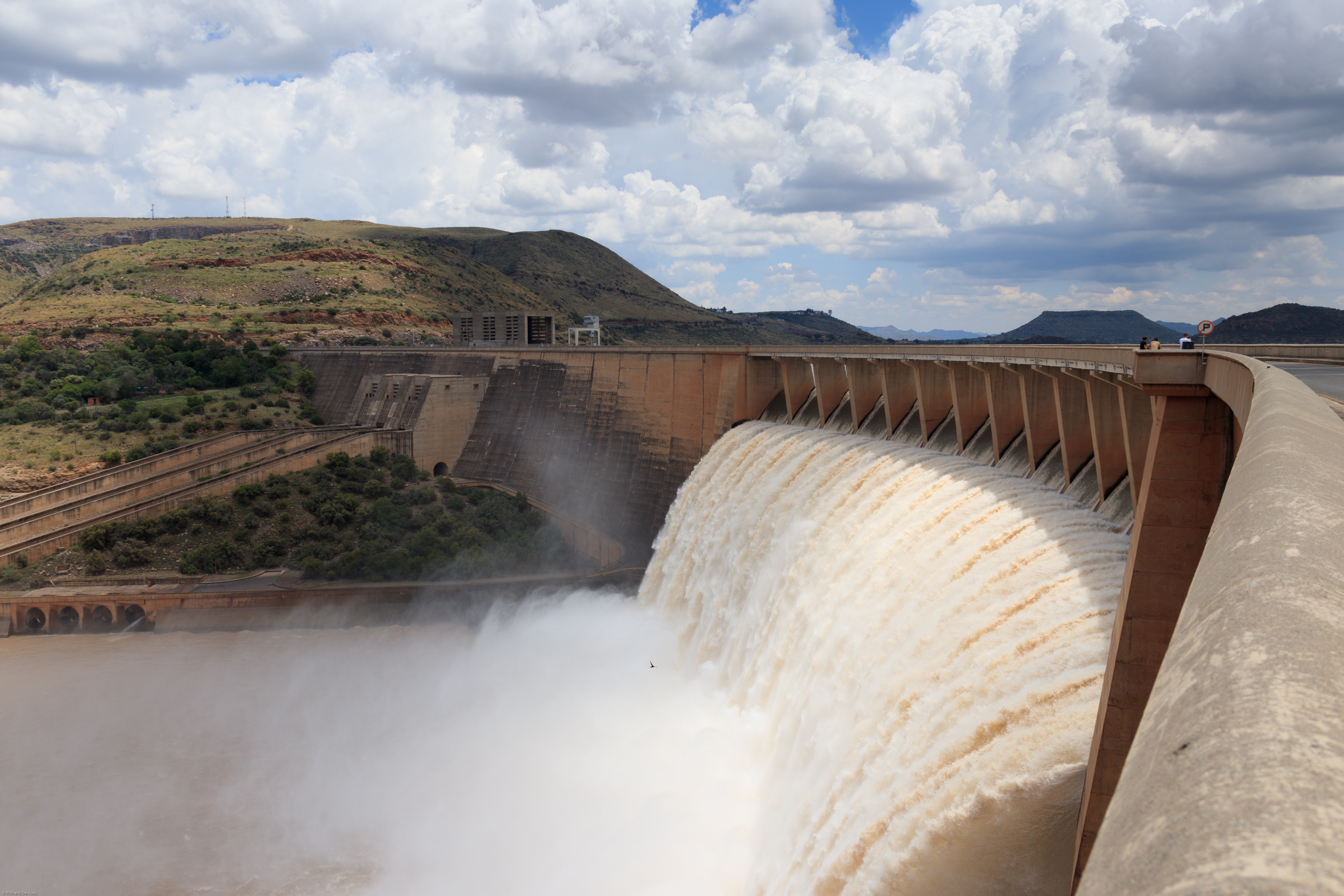 A large dam releasing a vast amount of water, creating mist. The structure stretches across a canyon with a partly cloudy sky and hills in the background.