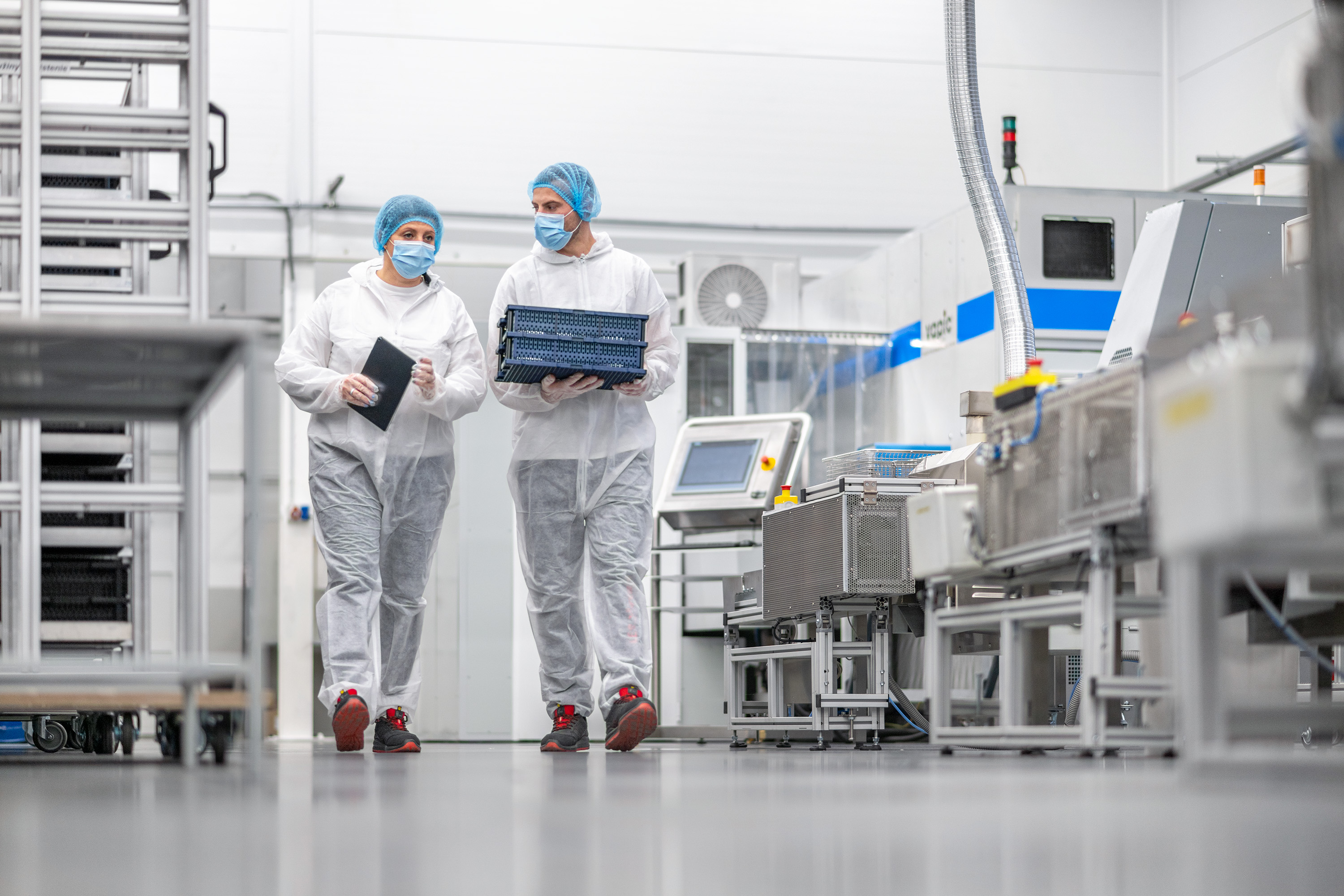 Two workers in cleanroom suits carrying a small crate through a sterile manufacturing environment.