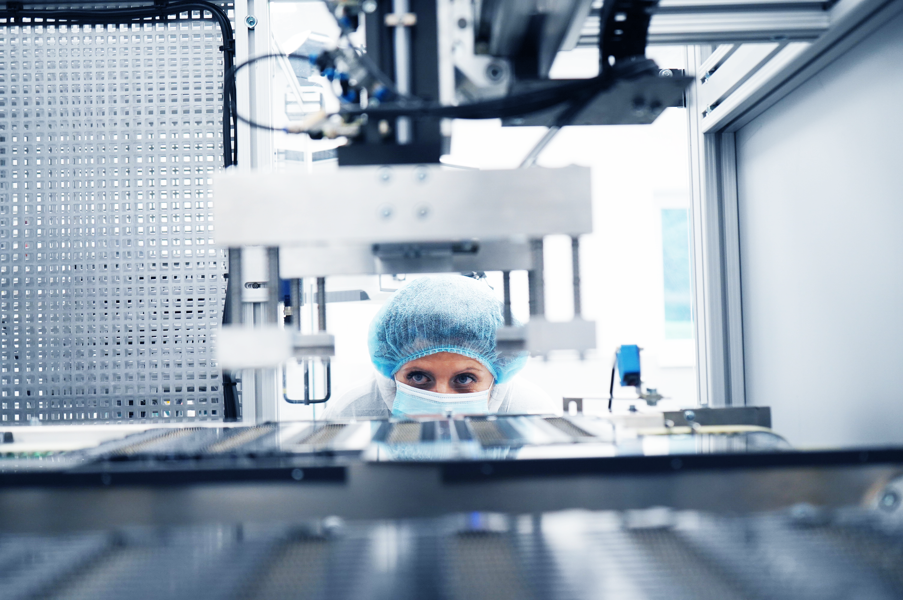 A worker wearing a hairnet and face mask closely inspecting automated machinery in a clean production setting.
