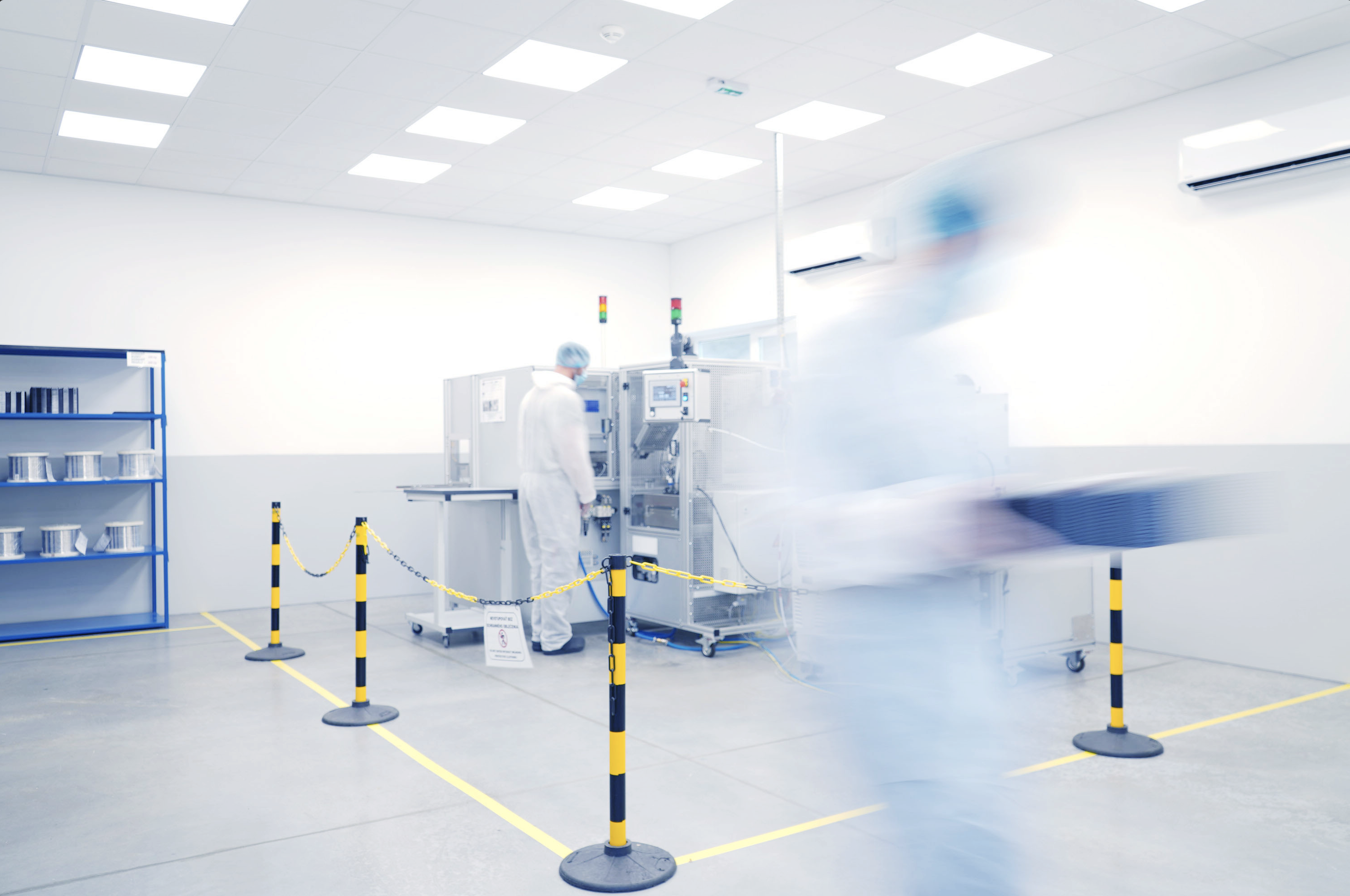 A blurred figure in a cleanroom suit moves quickly in a sterile laboratory. The room is bright with fluorescent lights, equipment, and safety barriers.