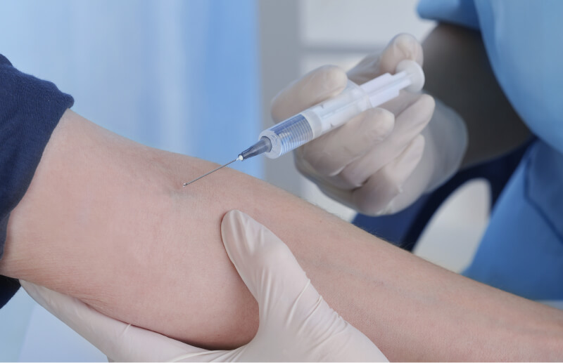 A healthcare professional in blue scrubs administers an injection into a patient's arm. The professional wears gloves.