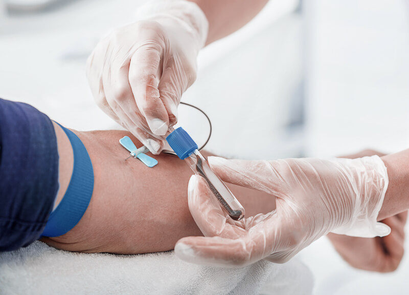 A healthcare professional with gloves draws blood from an arm. A blue tourniquet is tied around the arm, and a needle is inserted into a vein.