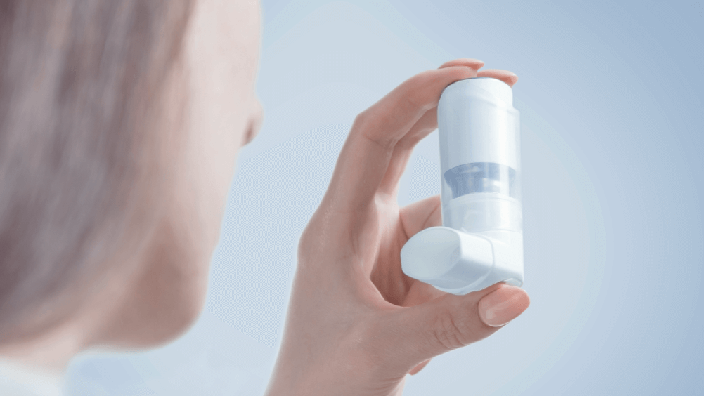 A person holds a white inhaler against a soft blue background. The focus is on the inhaler.