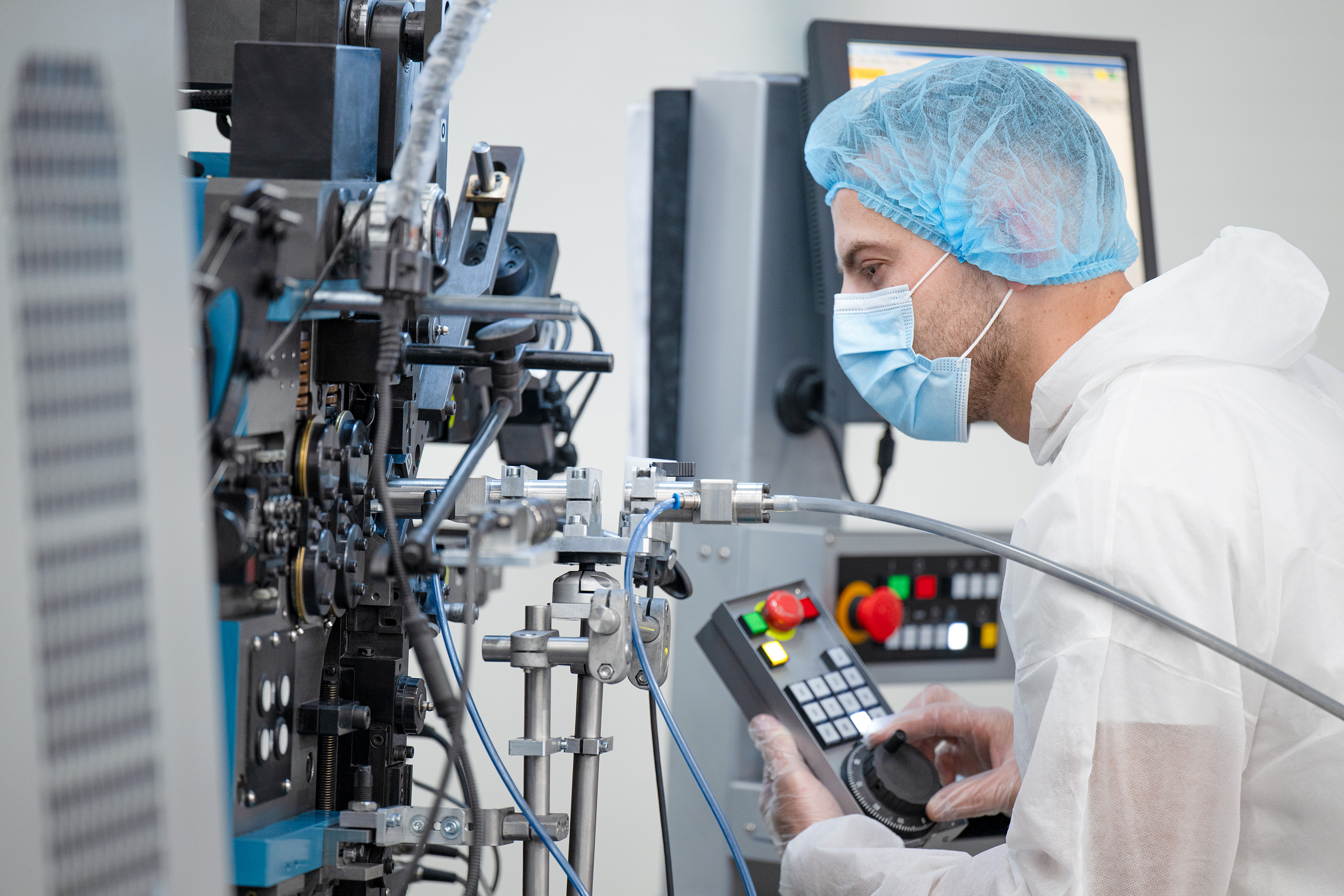 A technician in a white coat and blue hairnet operates a control panel in a high-tech lab. They focus on intricate machinery.