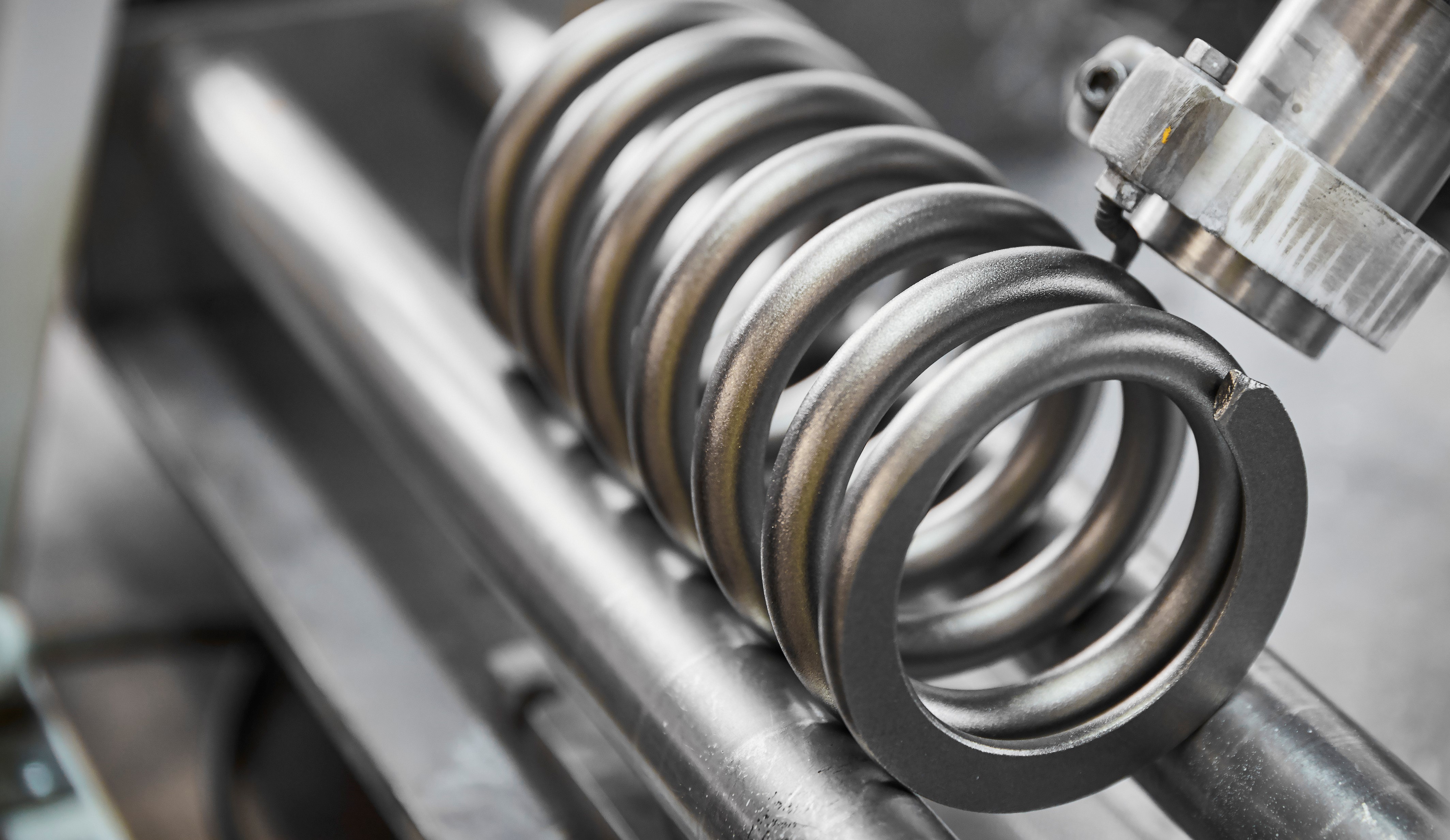 Close-up of a shiny metal compression spring on industrial machinery. The focus is on the spiral pattern, conveying precision and mechanical engineering.