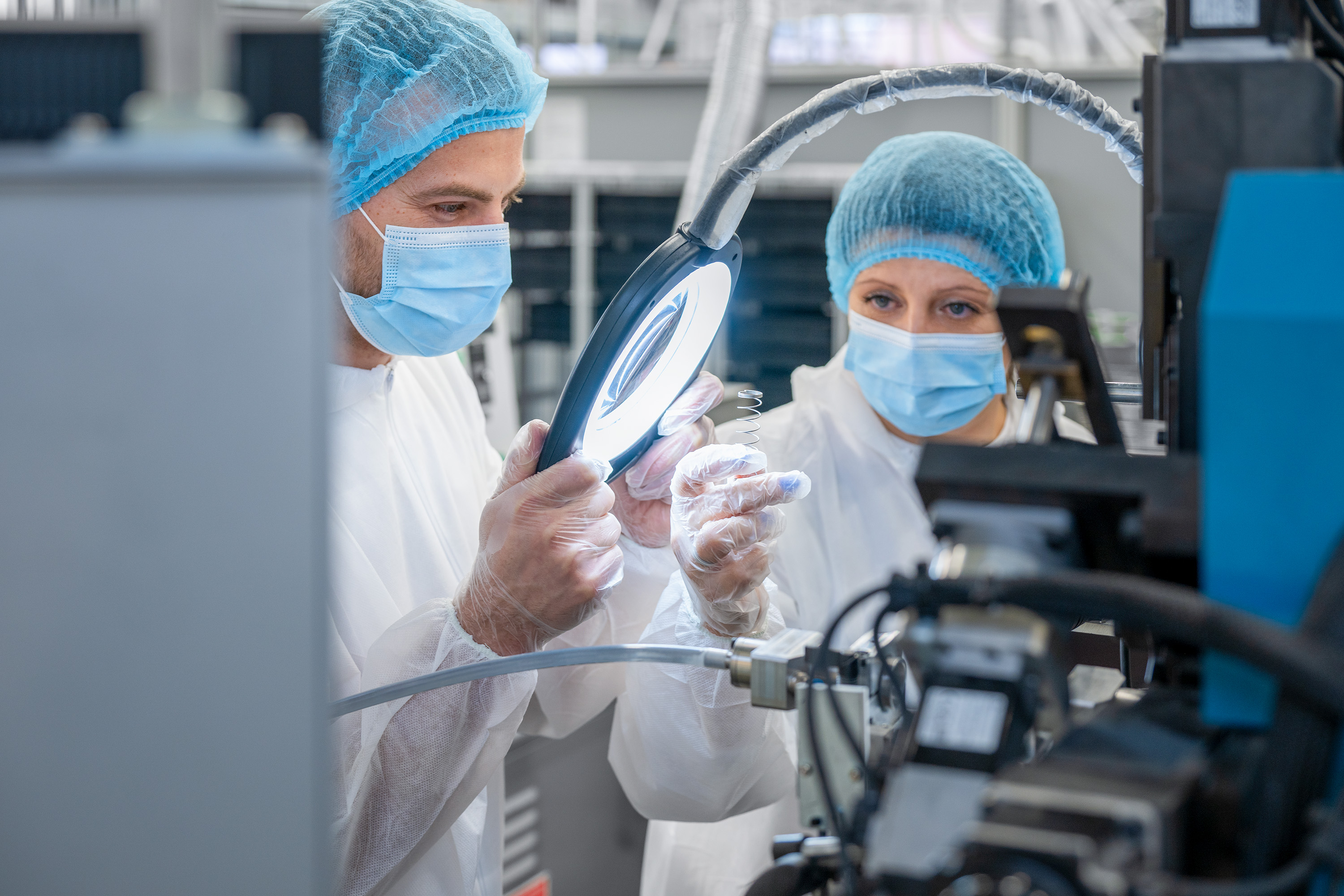 Two scientists in a lab, wearing masks and hairnets, examine a sample under a magnifying glass.