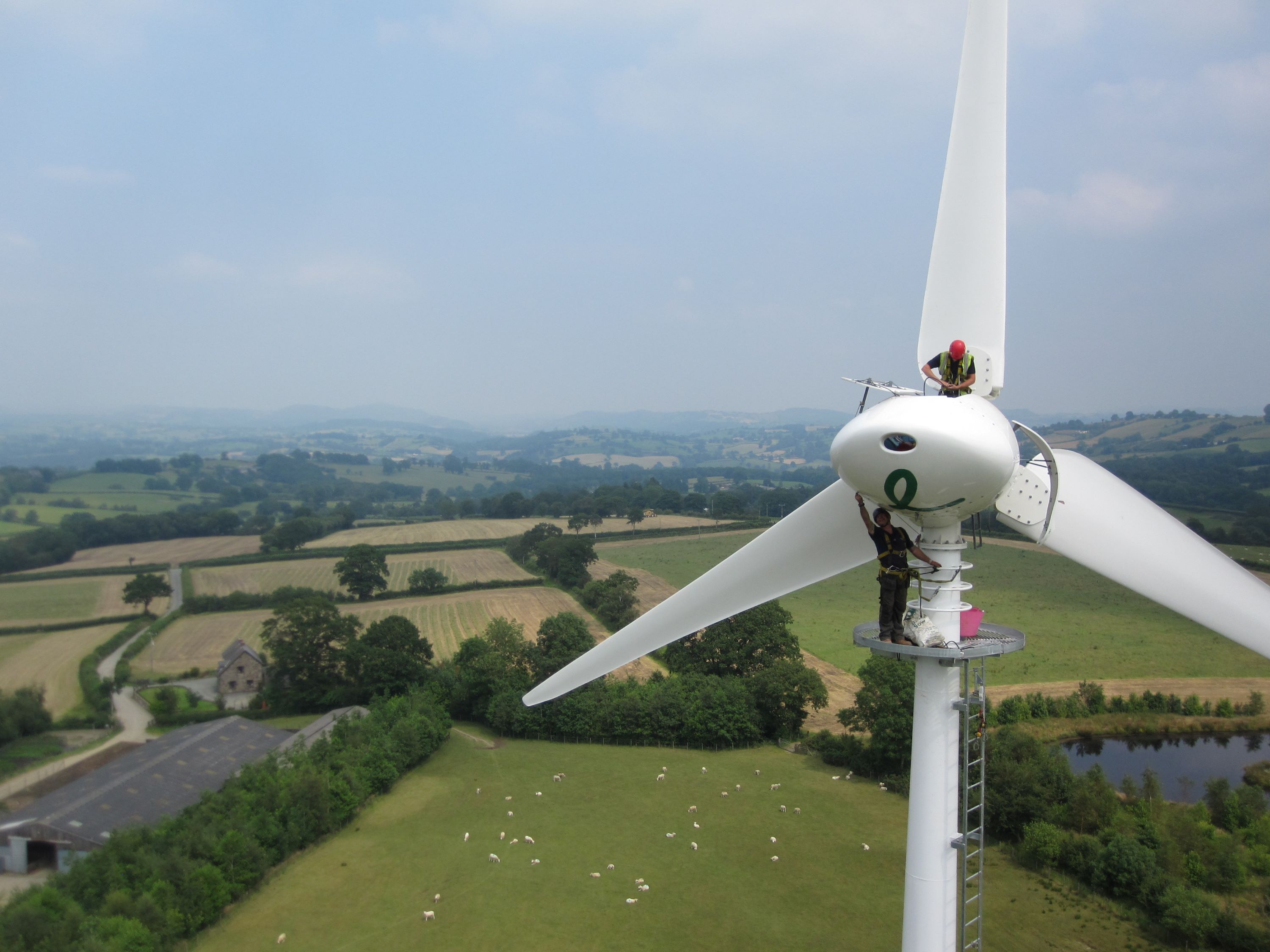 A person performs maintenance on a wind turbine in a vast, scenic countryside. The landscape features green fields and a cloudy blue sky.