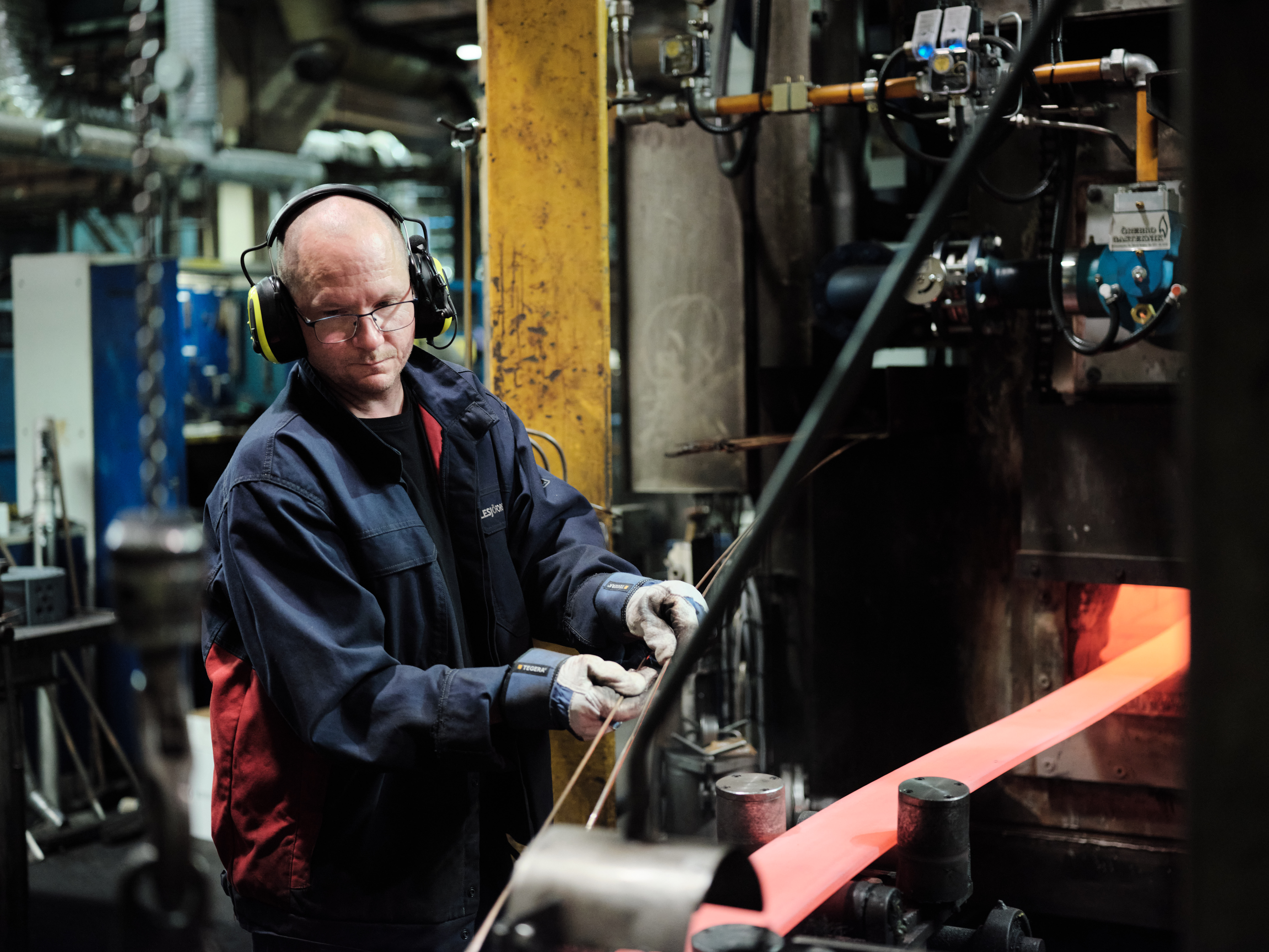 A worker in a workshop is shaping a glowing metal rod using heavy machinery. He wears ear protection and stands amidst industrial tools, focused on his task.