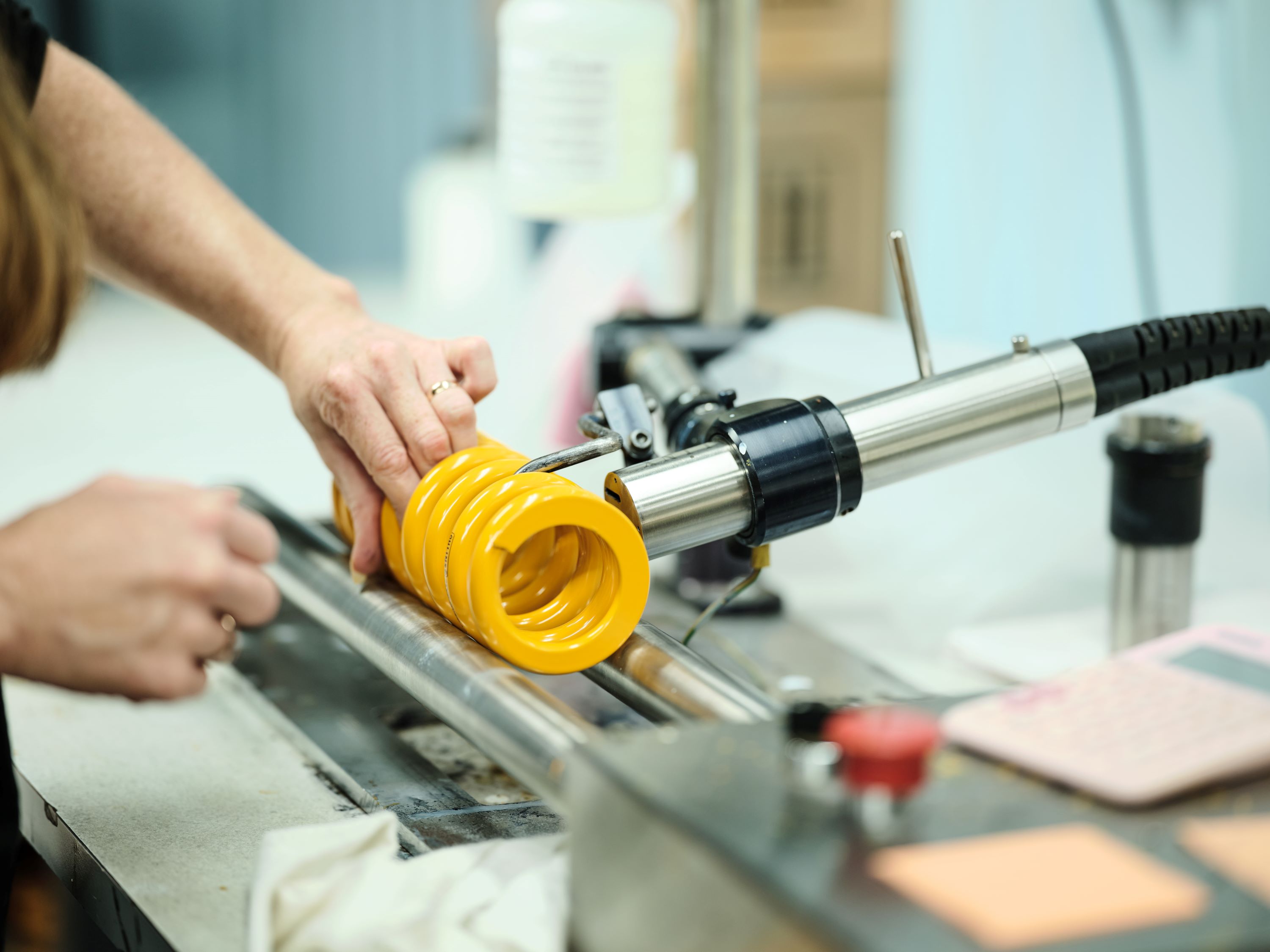 A person operates a machine winding a bright yellow spring onto a metal rod, The scene is industrial.