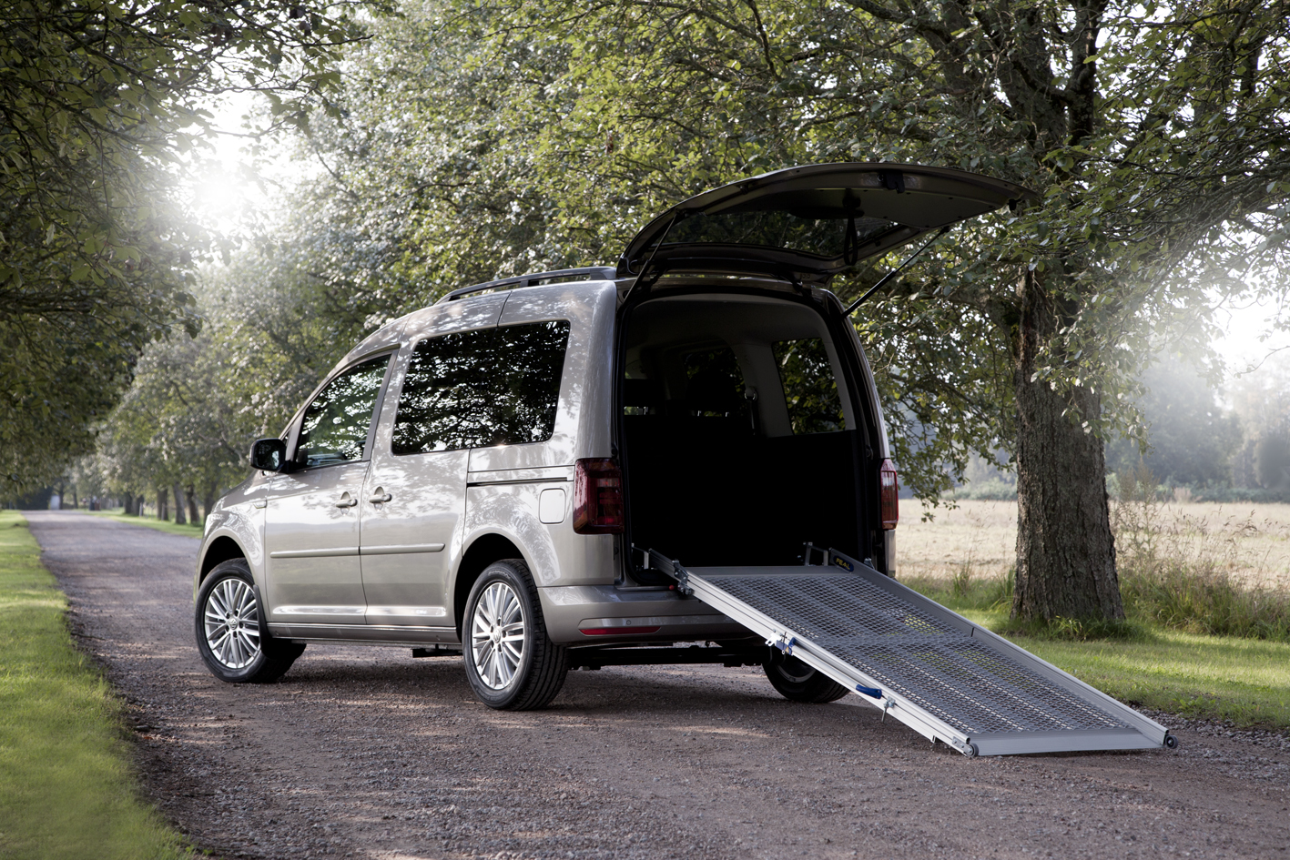 Silver van parked on a gravel road with a rear wheelchair access ramp extended.
