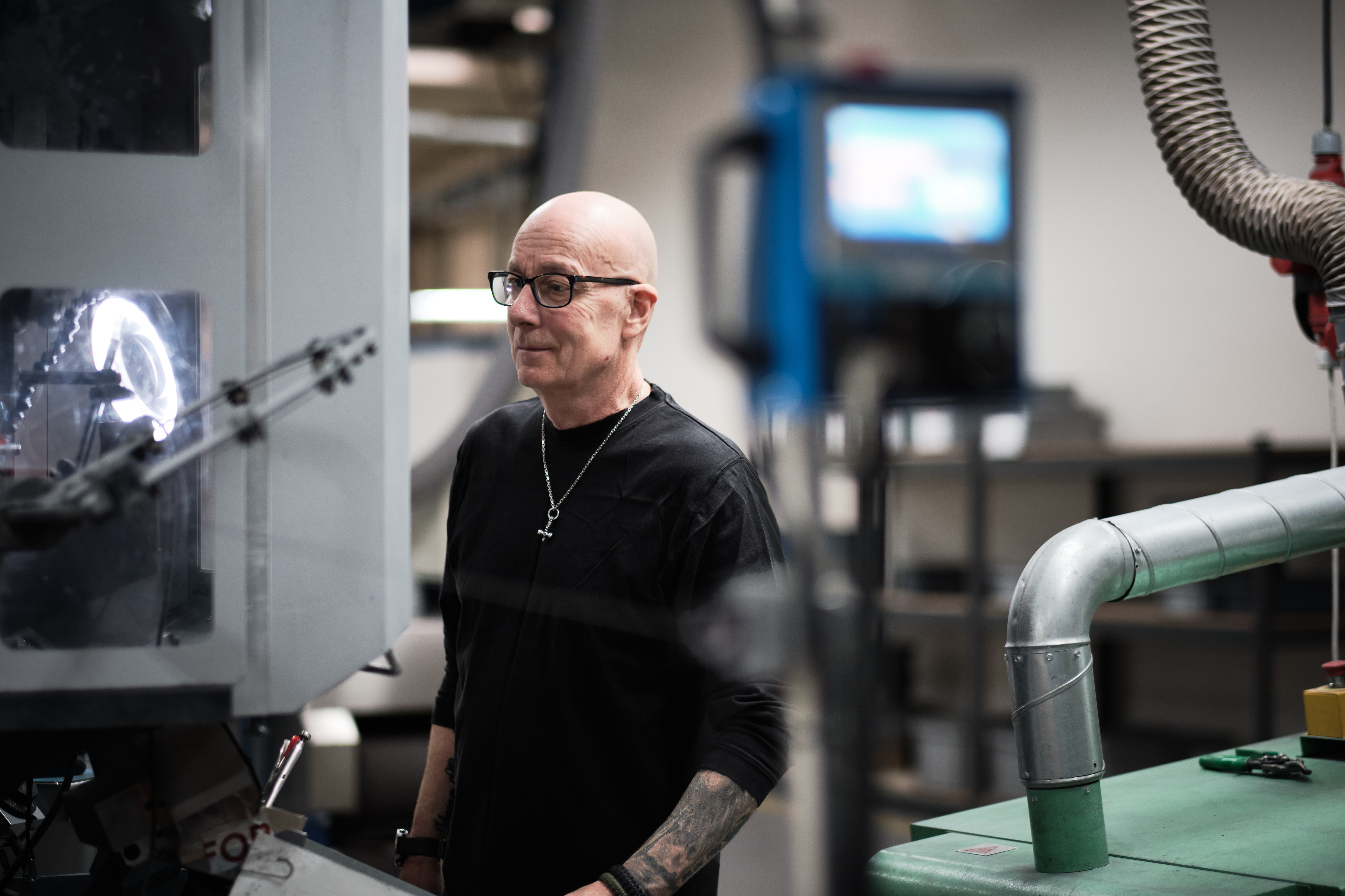 Man with glasses stands in a workshop, surrounded by machinery. He appears focused and engaged.