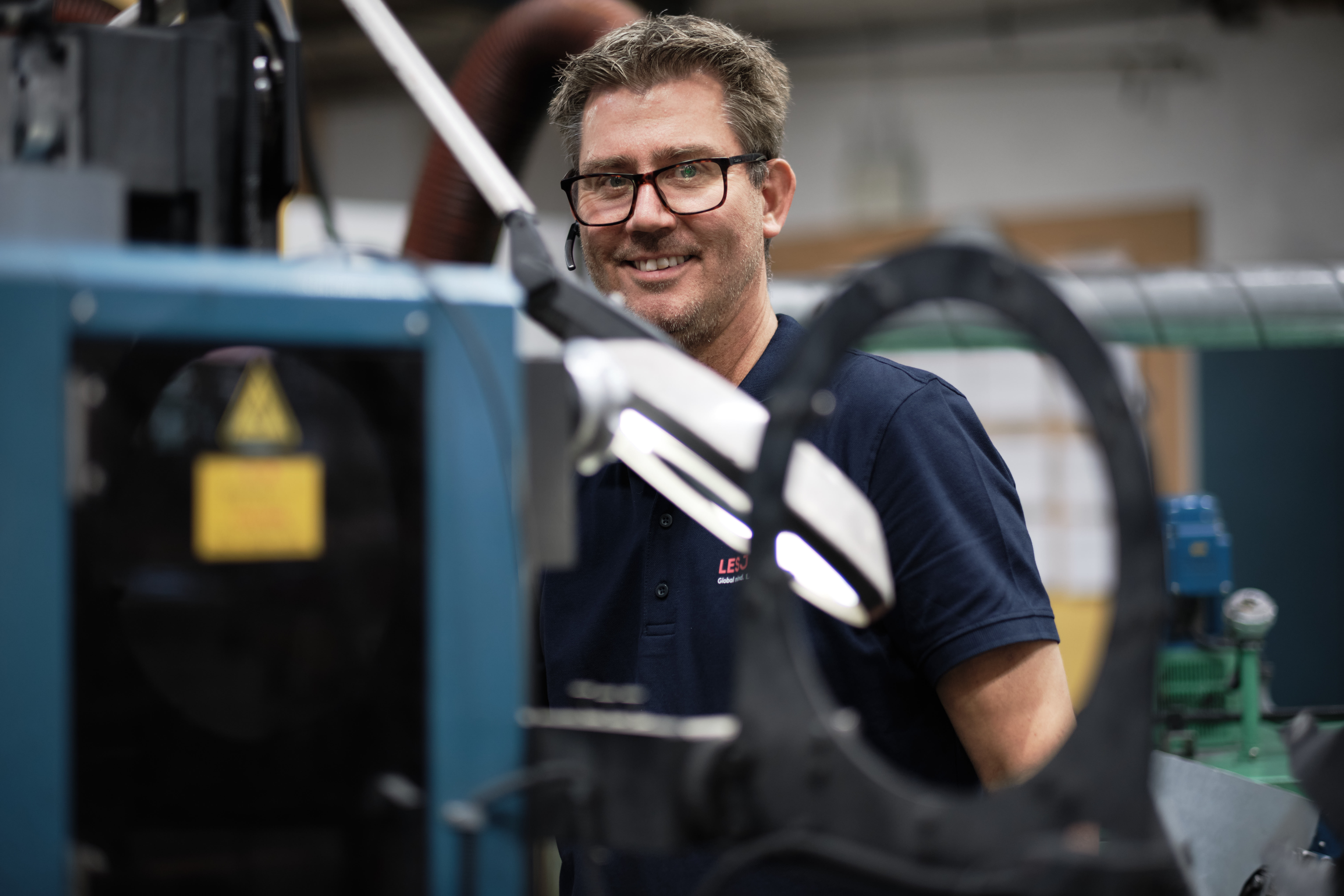 A smiling person in glasses and a blue shirt stands in a workshop surrounded by machinery, conveying a sense of skill and satisfaction.