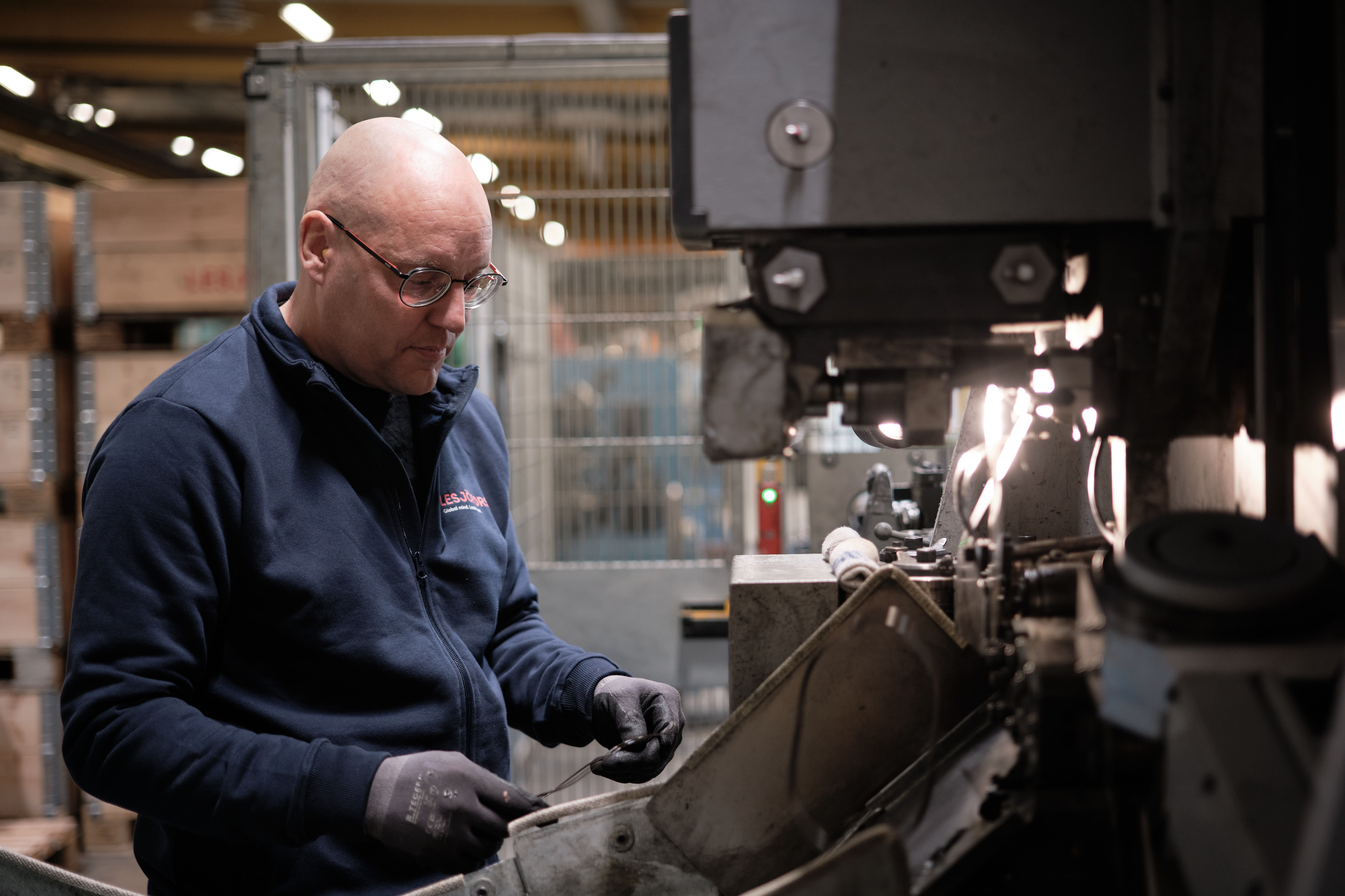 A man in a workshop carefully inspects a metal object under focused lighting, surrounded by machinery.
