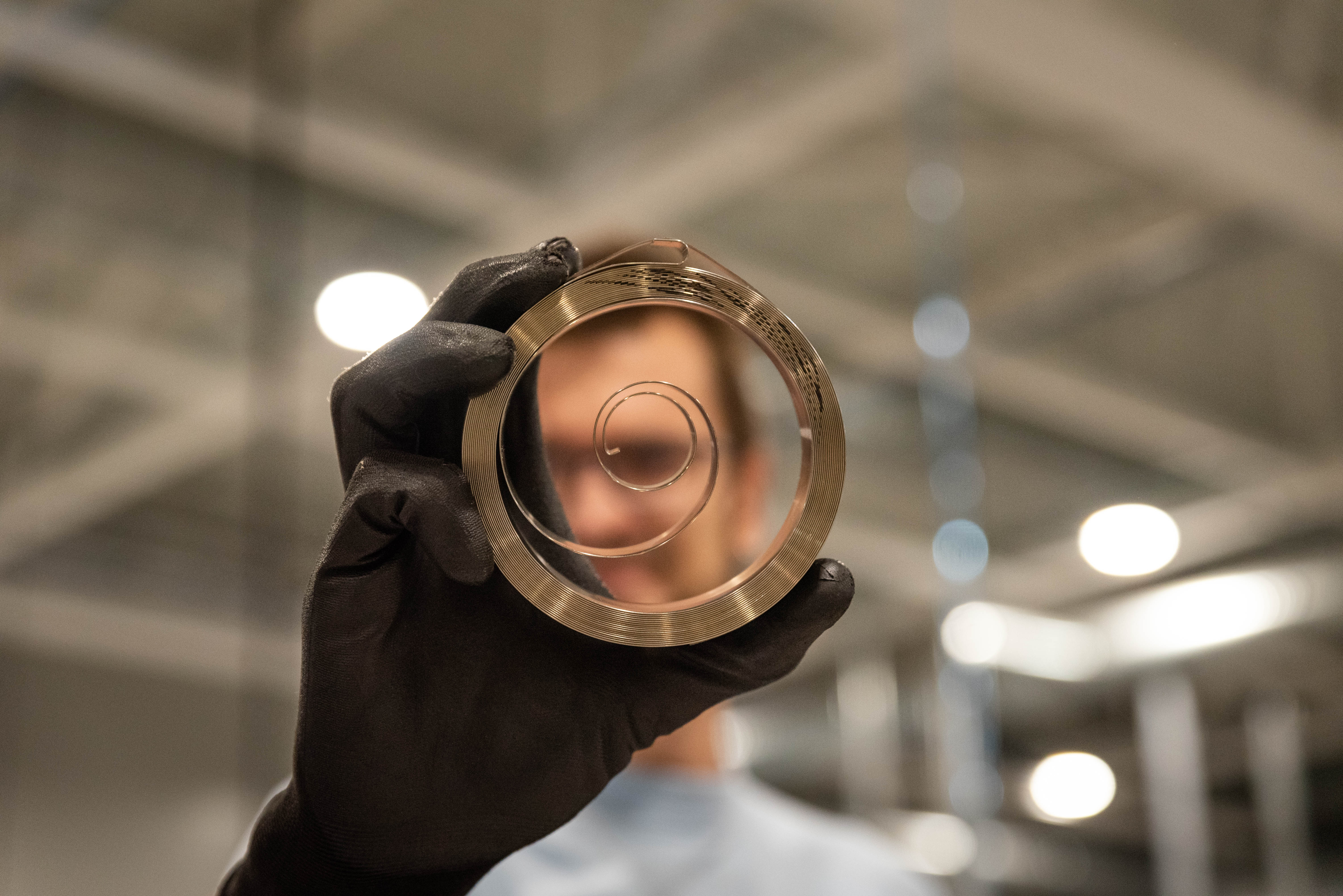 A person wearing black gloves holds up a flat spring, distorting their face behind it. The background is an industrial setting with blurred lights.