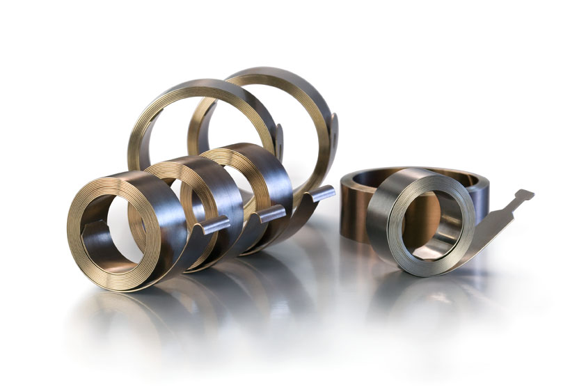 Five metallic flat springs of varying sizes are placed in a row on a reflective surface. The background is a soft white, creating a polished, industrial look.