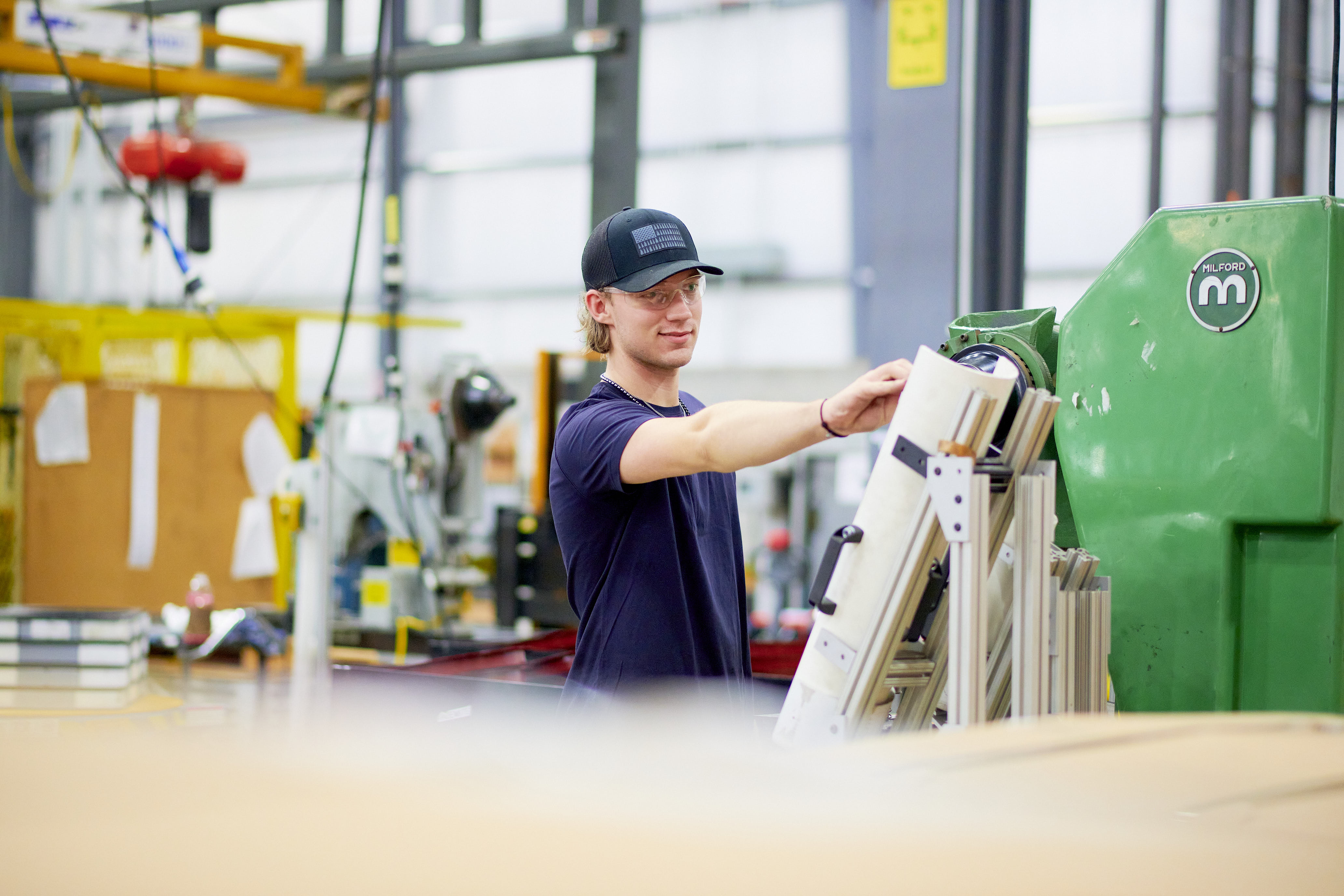 A worker in a factory, wearing a cap and navy shirt, operates machinery. The setting is industrial, with tools and equipment visible in the background.