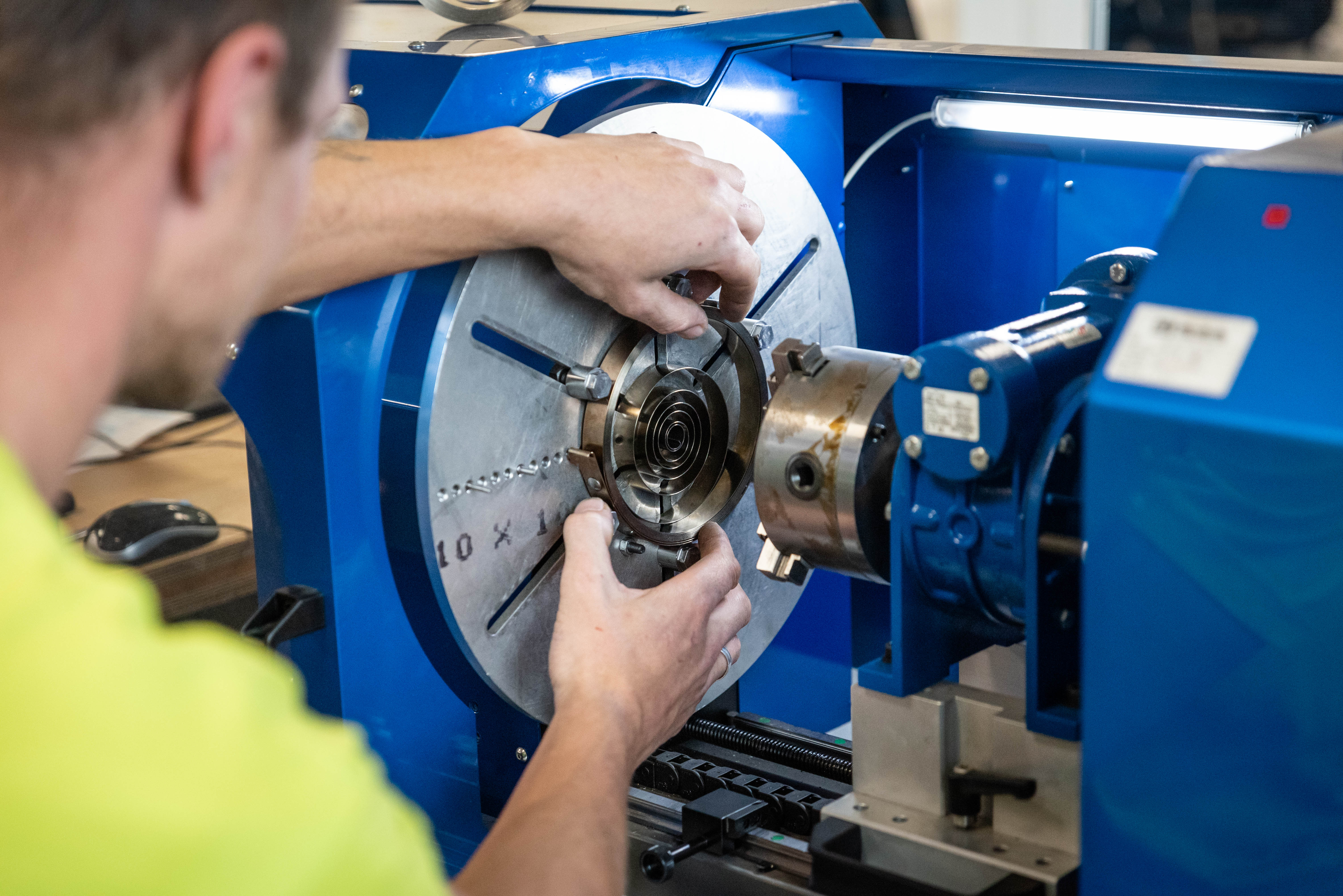 A person in a yellow shirt operates a blue machine, adjusting a mechanical part with precision, highlighting industrial focus and concentration.