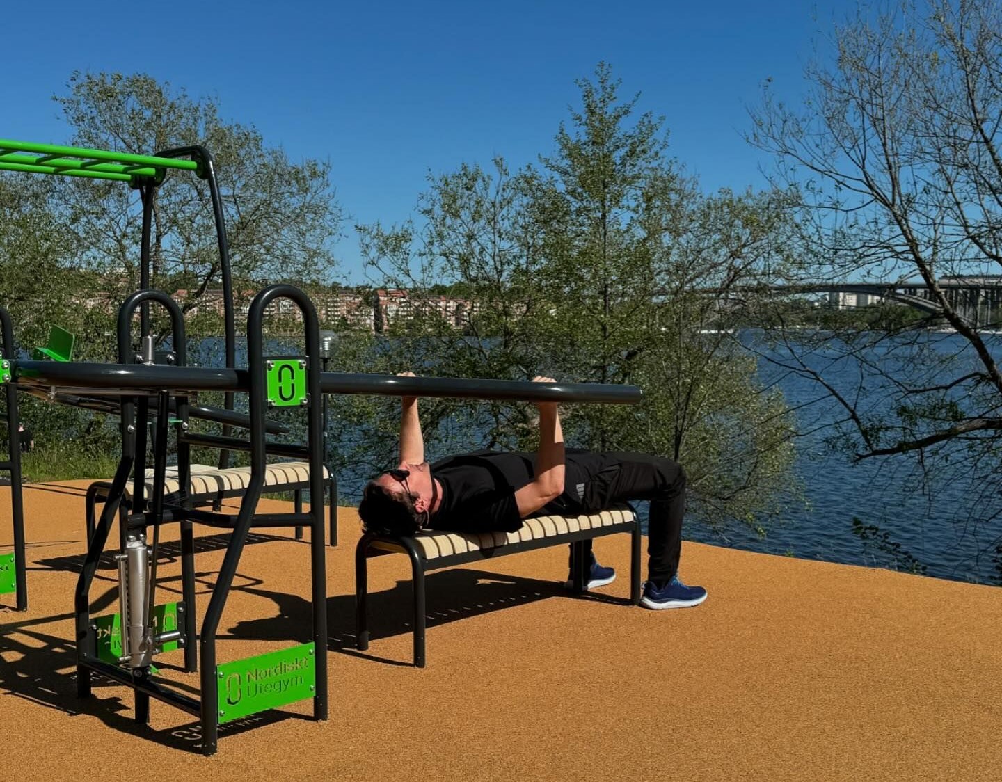 A person exercises on outdoor gym equipment by a river, under clear blue skies. Trees and a distant bridge create a peaceful backdrop.