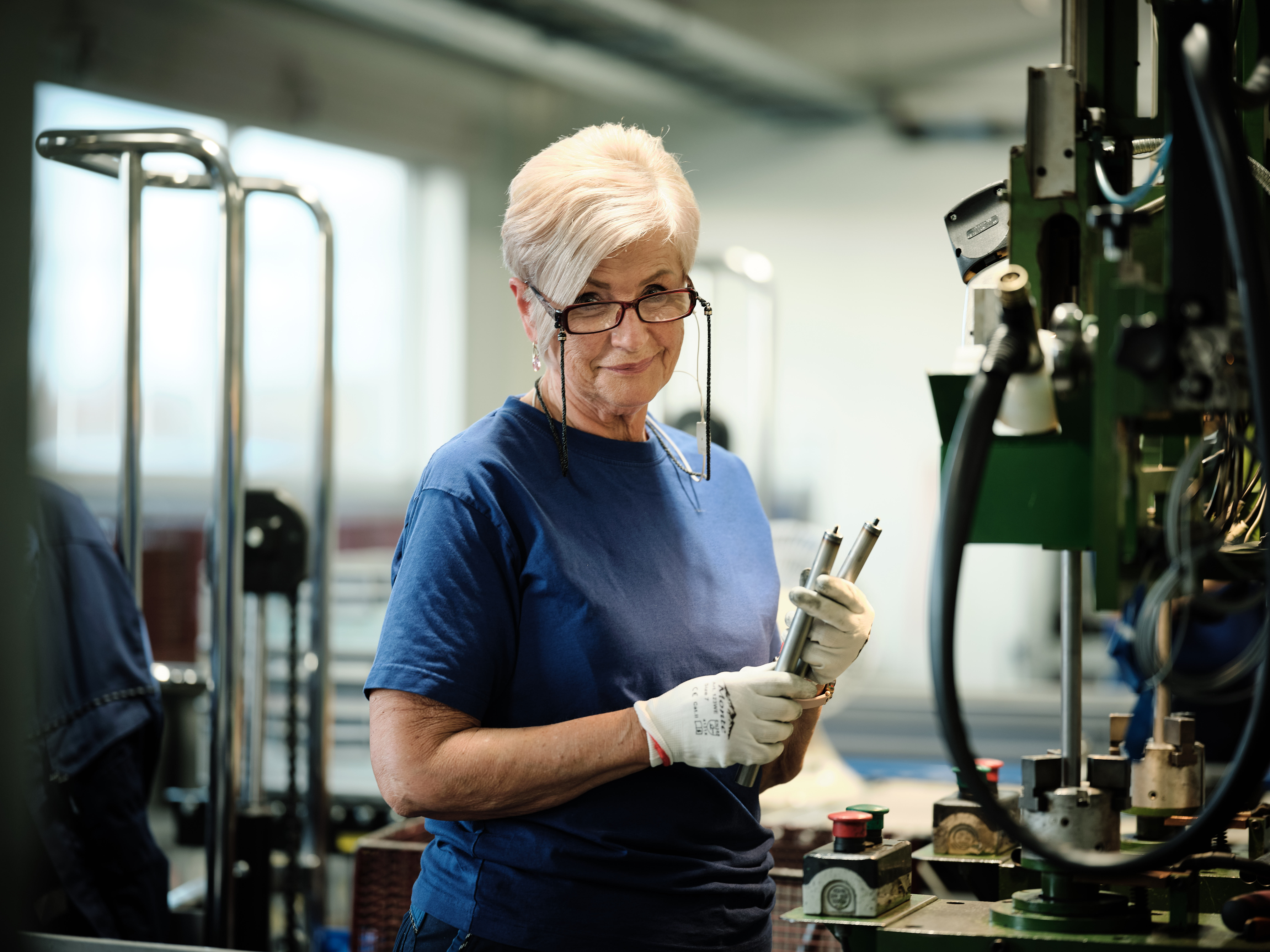 A women with glasses works in a factory, inspecting a metal part. She wears a blue shirt and gloves, focused and attentive. Machinery surrounds them.
