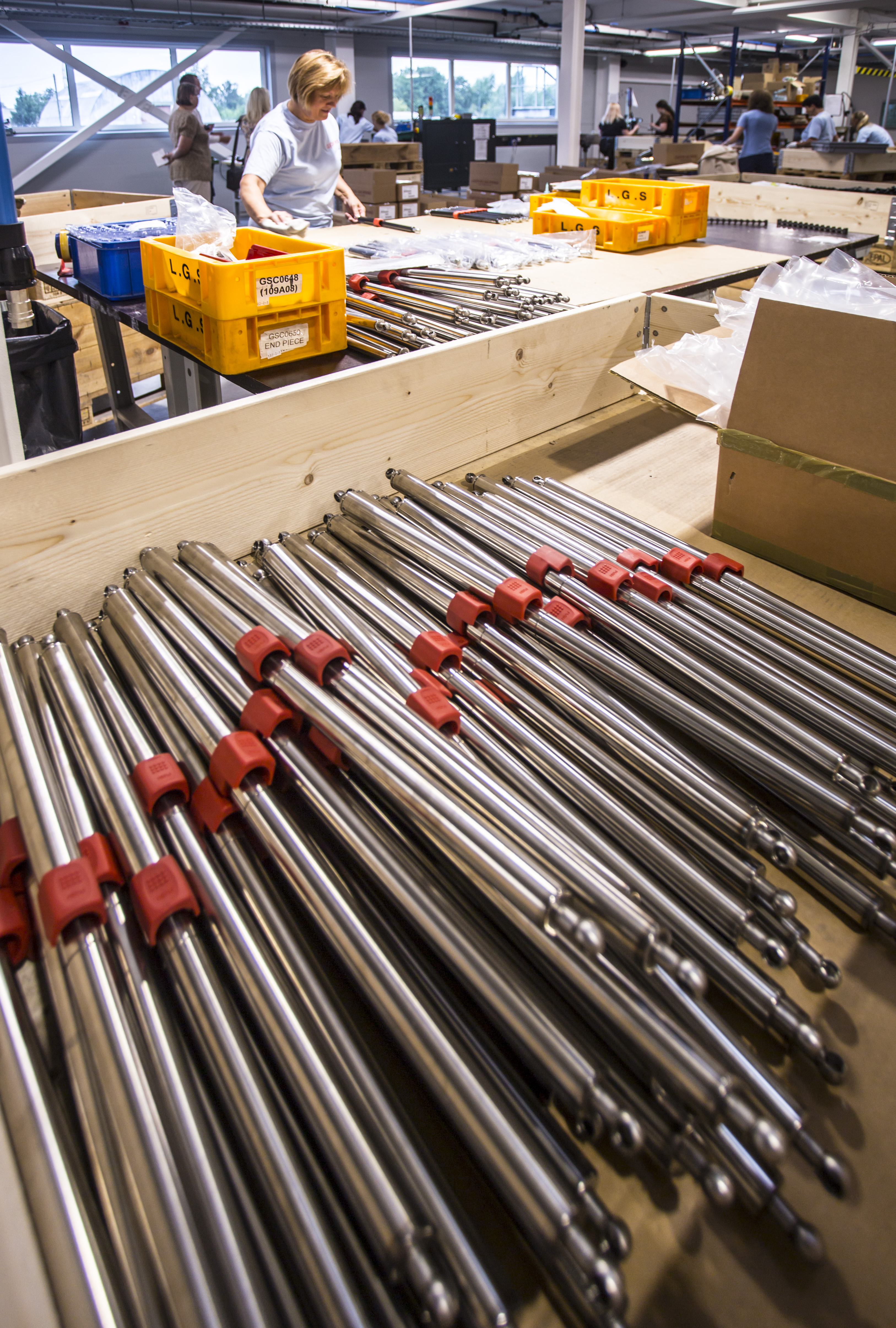 Stacks of long metal lockable springs with red caps are arranged on a factory floor in a wooden box.