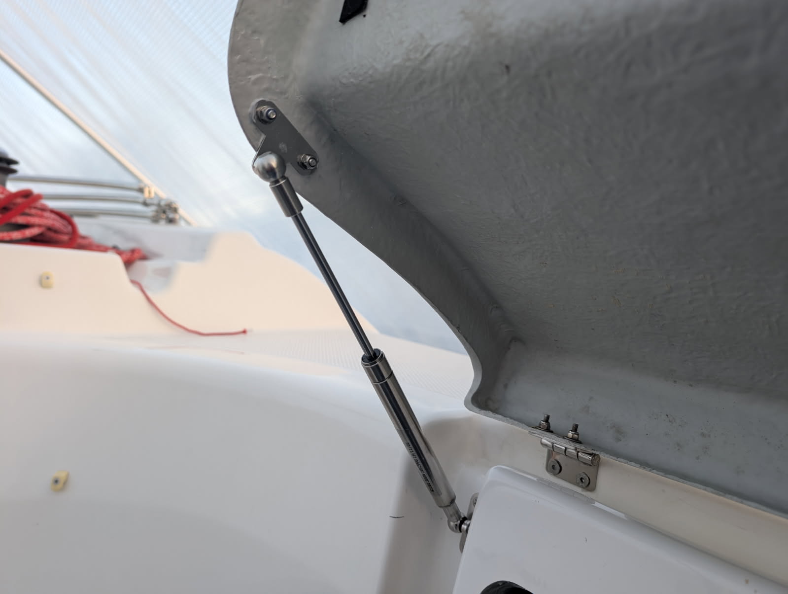 Close-up of a boat's open hatch with a gas spring. The background shows clear sky and rigging.
