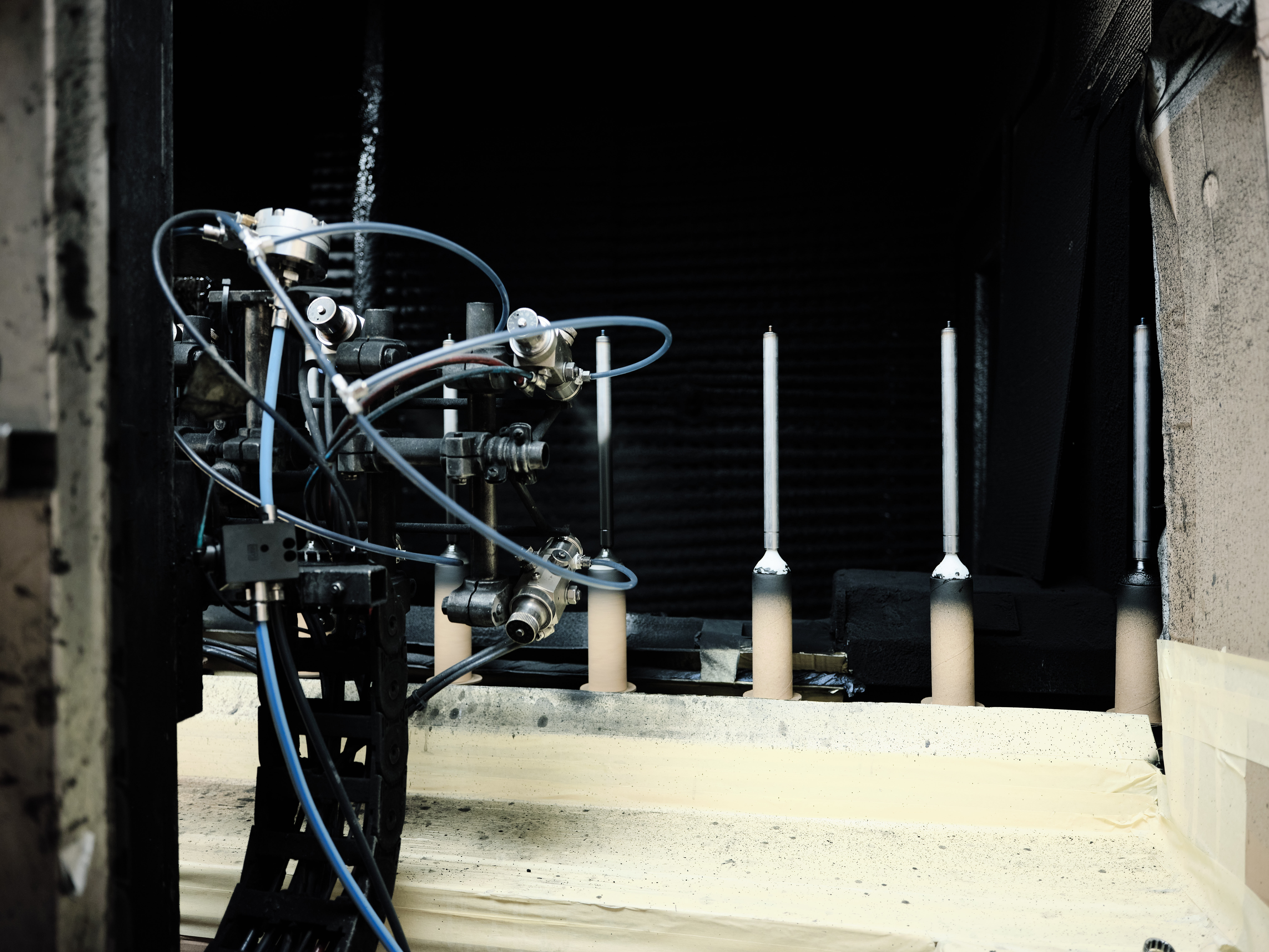 A complex robotic arm with tubes and wires is positioned in a dimly lit industrial setting. Four cylindrical rods are aligned in the background.