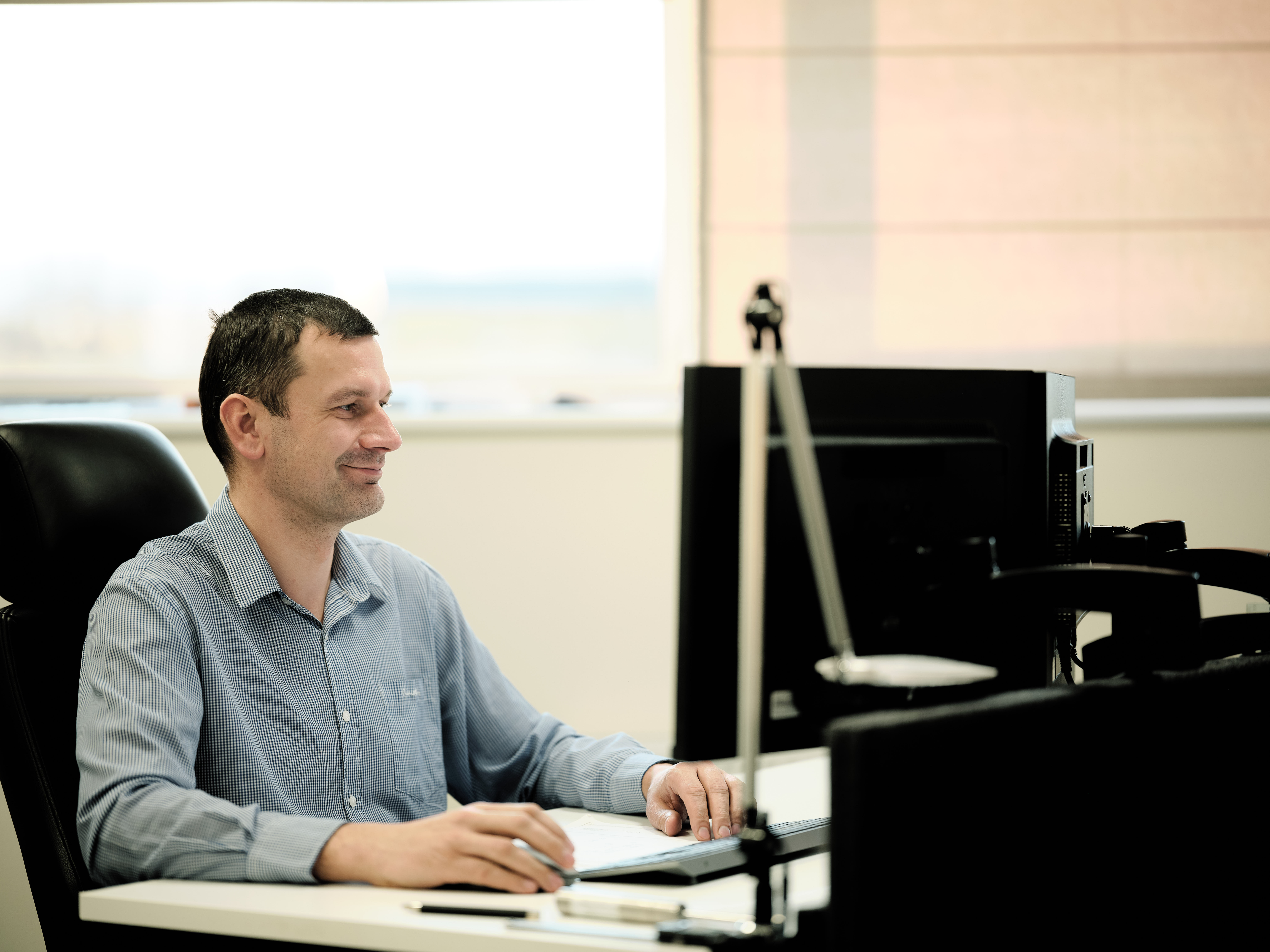 A person sits at a desk in an office, working on a computer. The scene is calm and focused, featuring soft lighting from a nearby window.