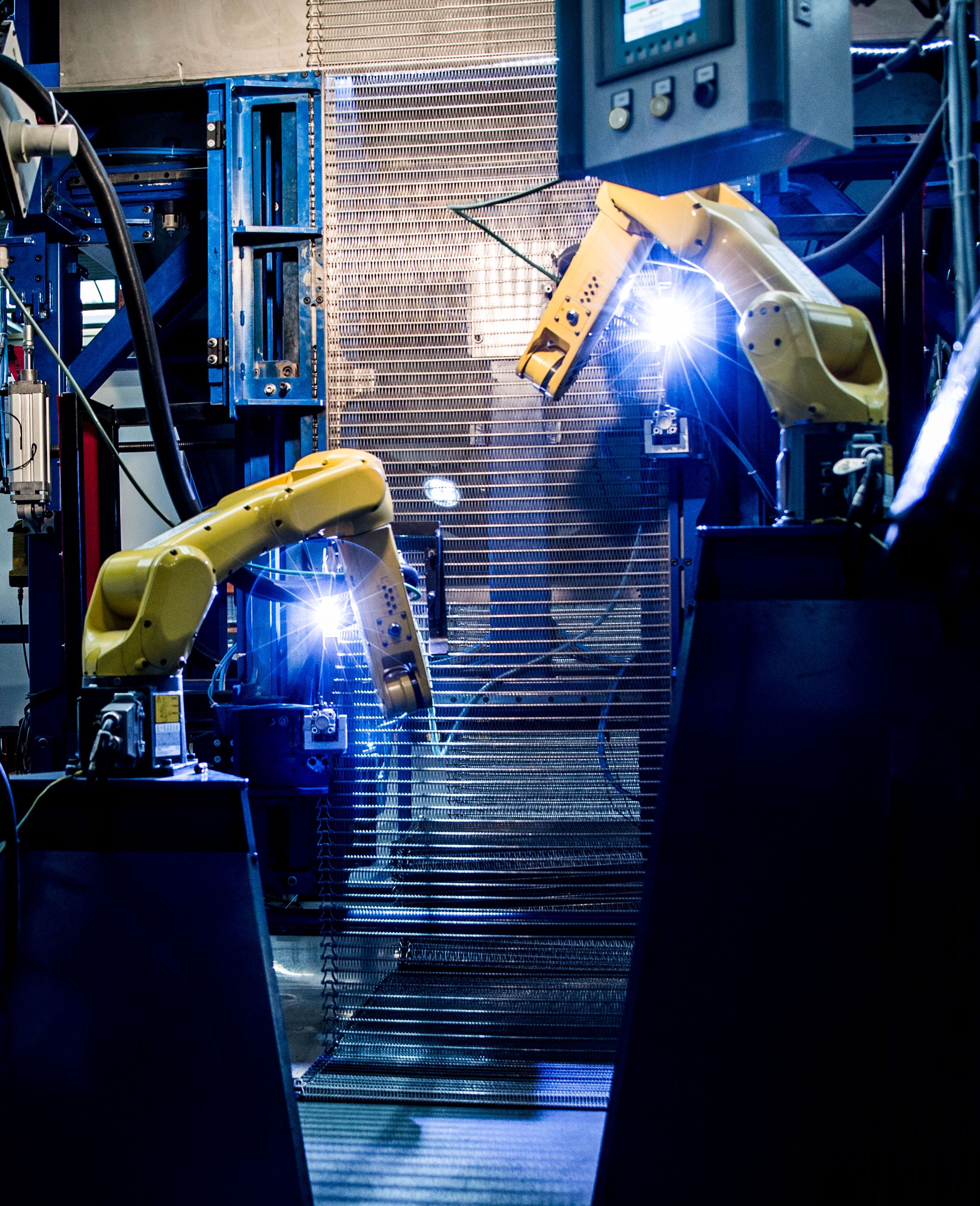 Two yellow robotic arms welding in a dimly lit factory. Blue sparks illuminate the industrial setting, highlighting advanced manufacturing technology.