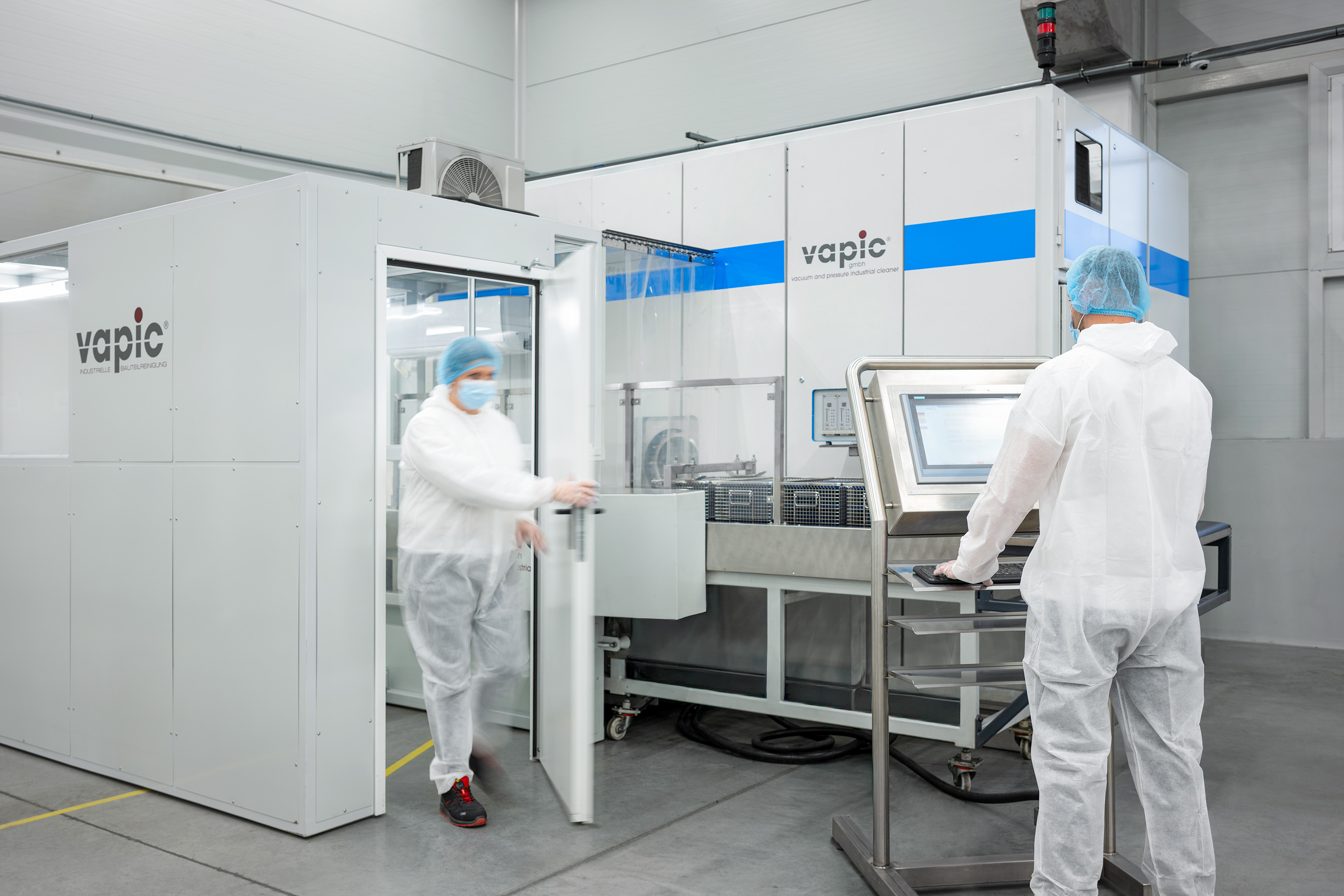 Two workers in cleanroom suits and blue caps operate machinery in a sterile lab.