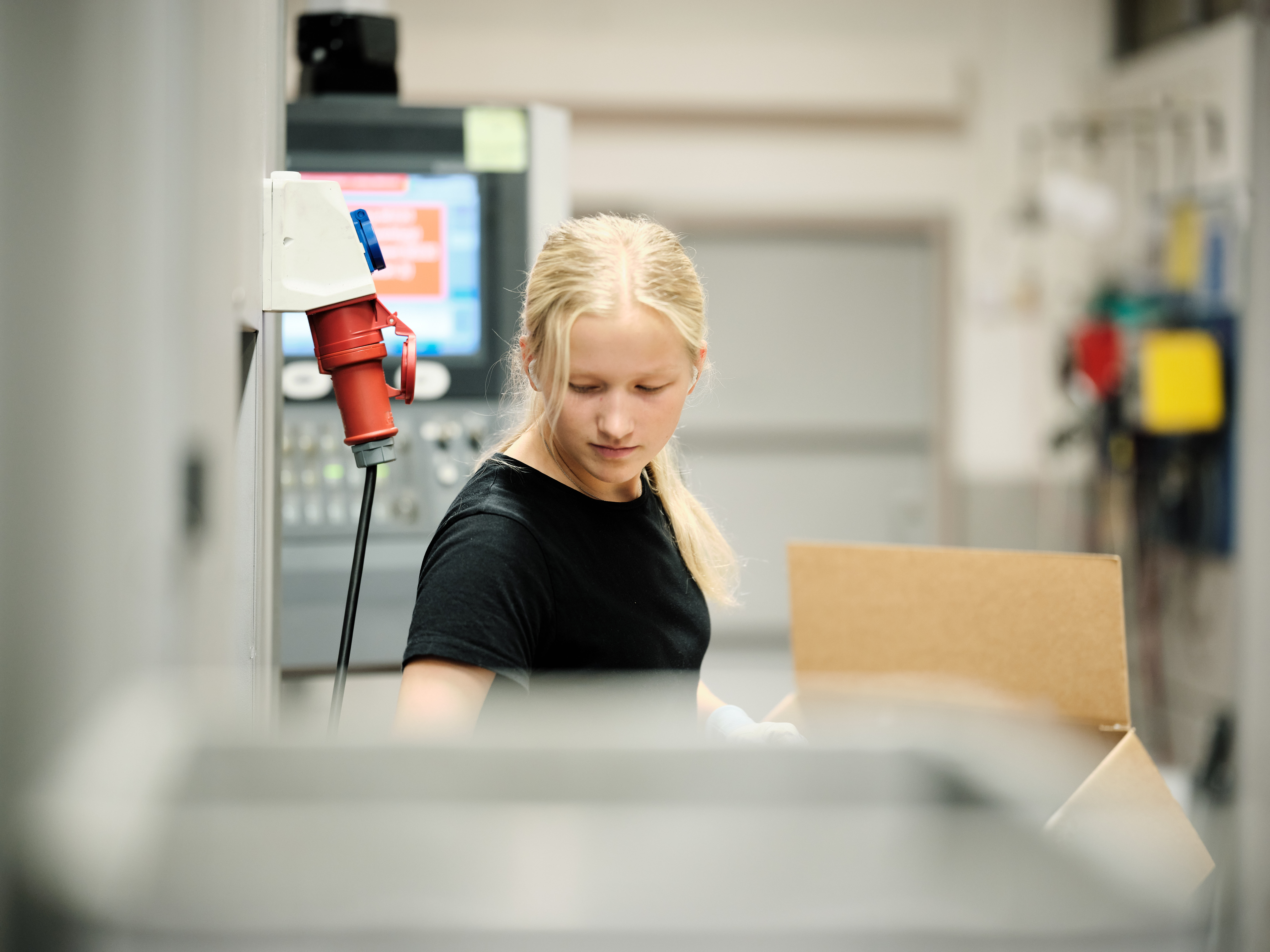 A factory worker holding a cardboard box, with machinery and control panels around her.