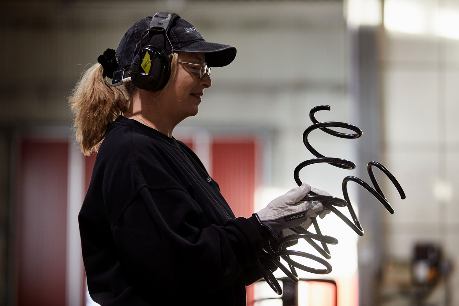 A worker wearing a cap, ear protection, and gloves holding and looking at a large black coil spring, with industrial equipment blurred in the background.