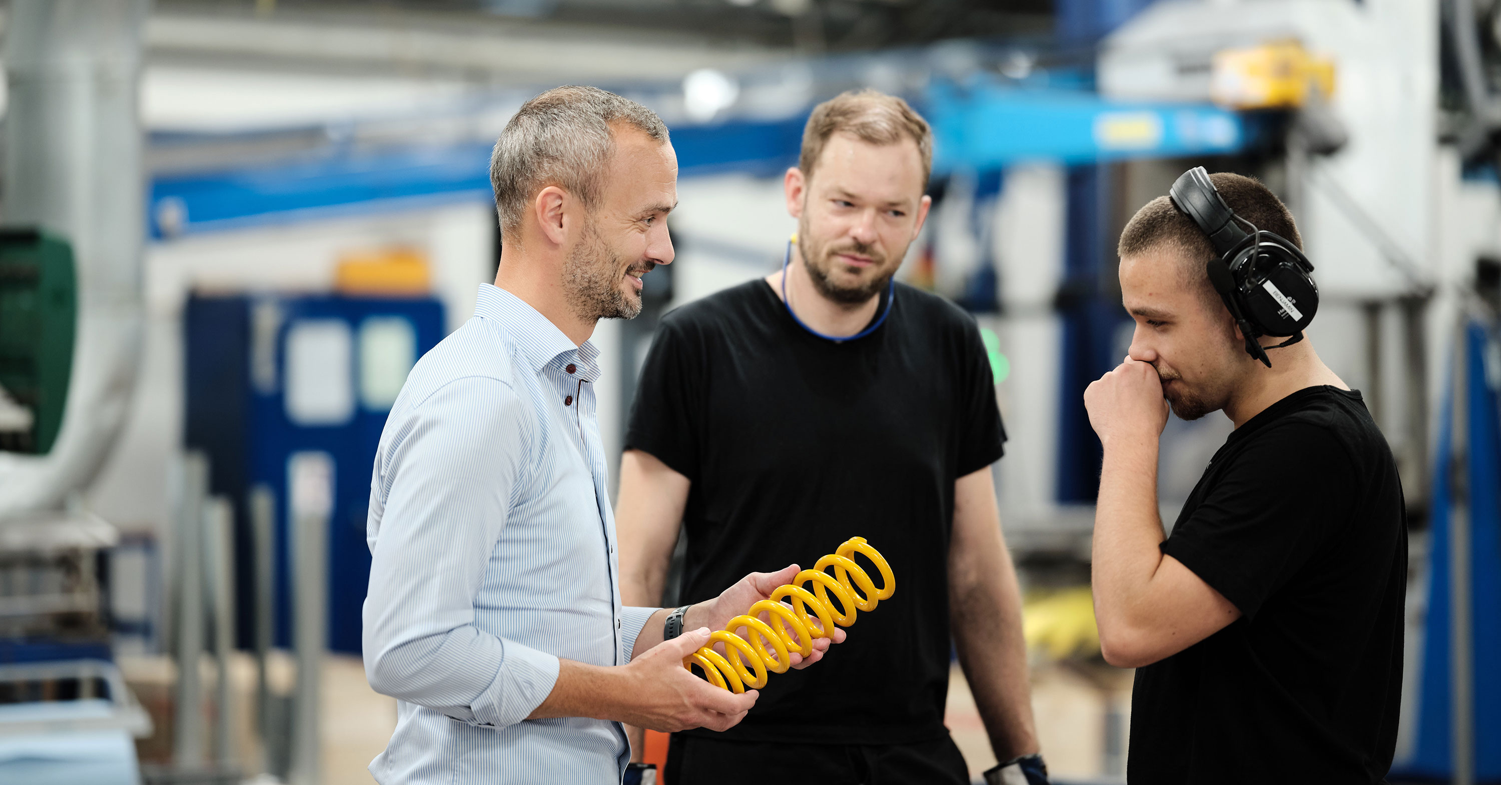Three men standing on a factory floor discuss a bright yellow coil spring, with machinery blurred in the background.